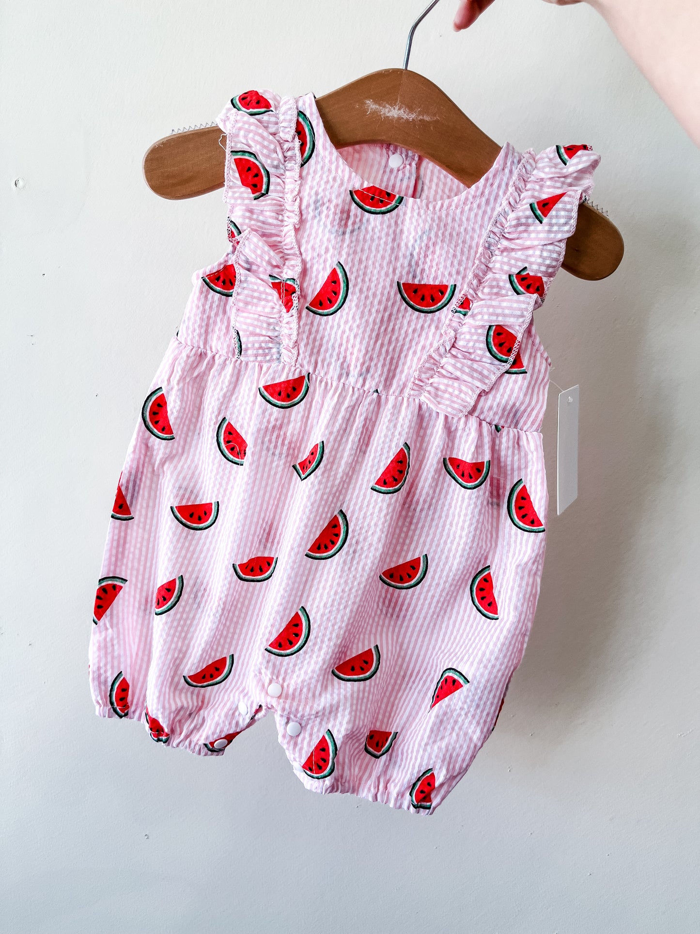 The Pink Picnic Watermelon Romper by Tiny Tot Bundles features ruffled sleeves and a playful watermelon print, displayed on a wooden hanger against a light wall.