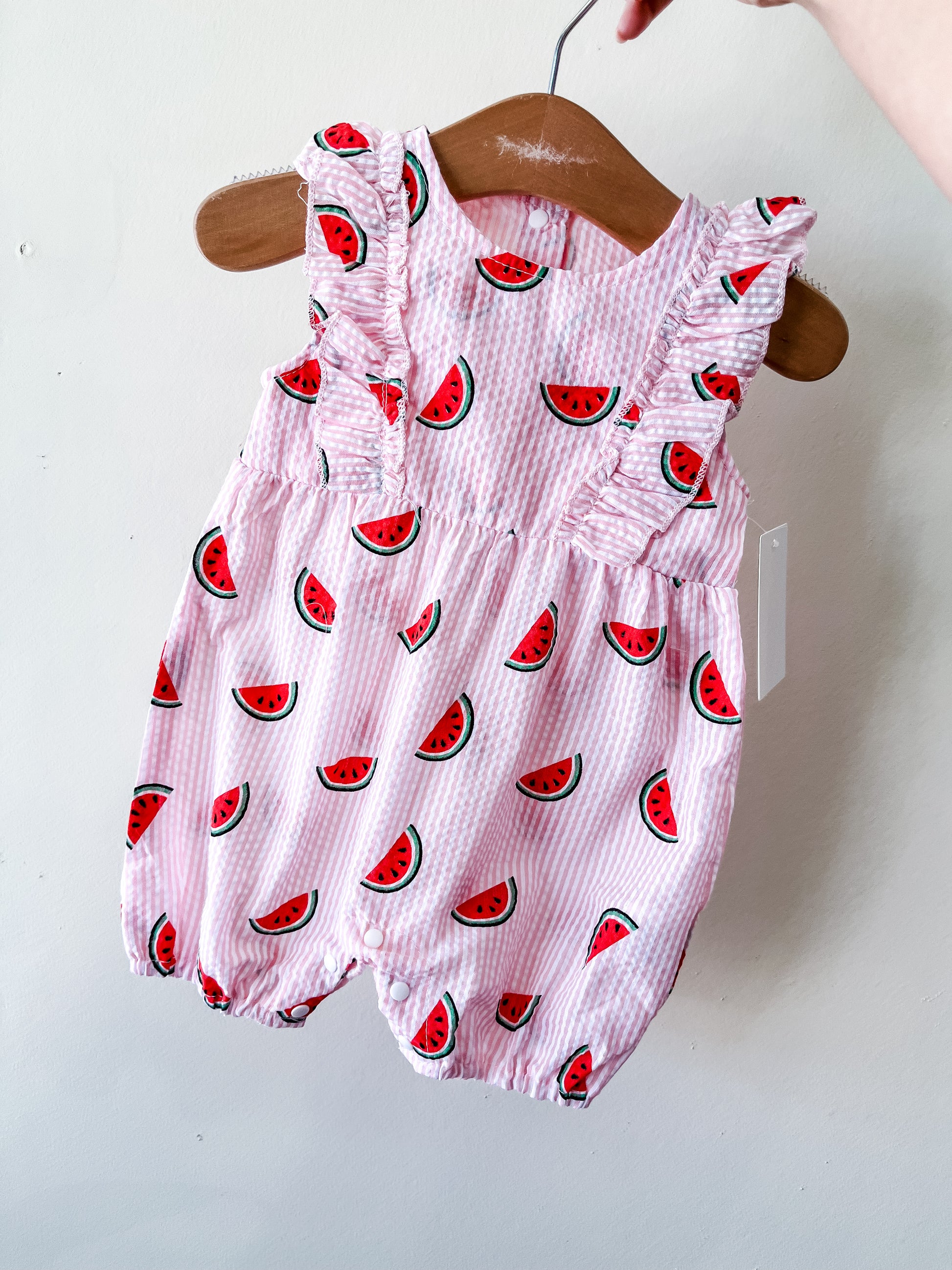 The Pink Picnic Watermelon Romper by Tiny Tot Bundles features ruffled sleeves and a playful watermelon print, displayed on a wooden hanger against a light wall.