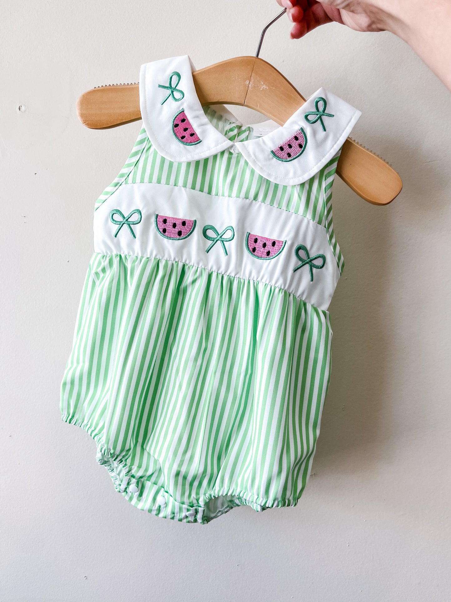 The Tiny Tot Bundles Watermelon Picnic Bubble is a sleeveless baby romper in green and white stripes with a white collar and chest, embroidered watermelon slices, green bows, and elastic leg openings, shown hanging on a wooden hanger.
