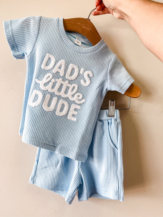 A hand displays Tiny Tot Bundles' Dad's Little Dude Set—a light blue ribbed short-sleeve shirt with "DADS little DUDE" in textured white letters and matching shorts—against a plain beige wall.