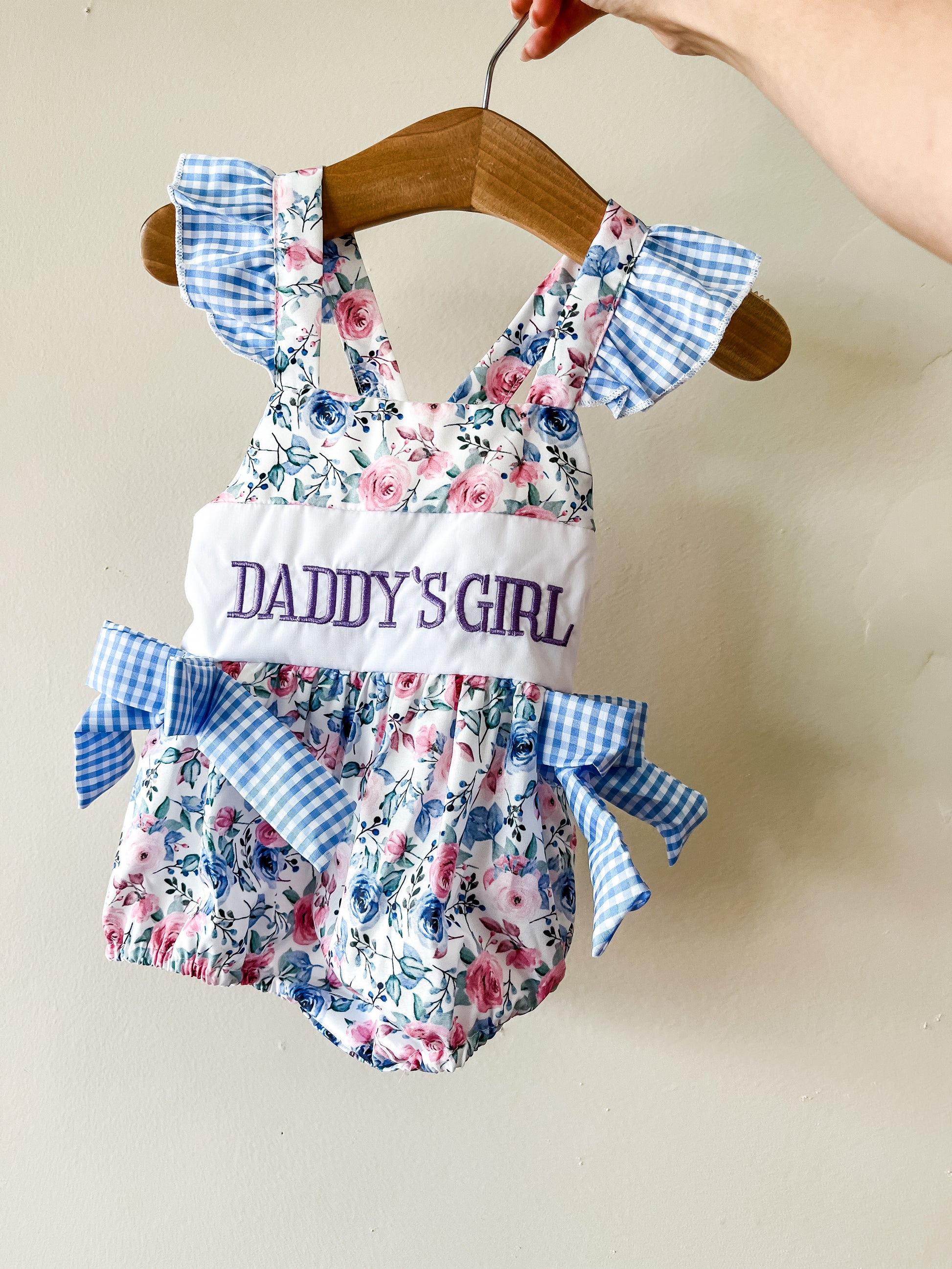 The Daddy’s Girl Floral Bubble by Tiny Tot Bundles features blue and pink flowers, ruffled blue gingham sleeves with bows, and “DADDYS GIRL” embroidered in purple across the chest. Shown on a wooden hanger.