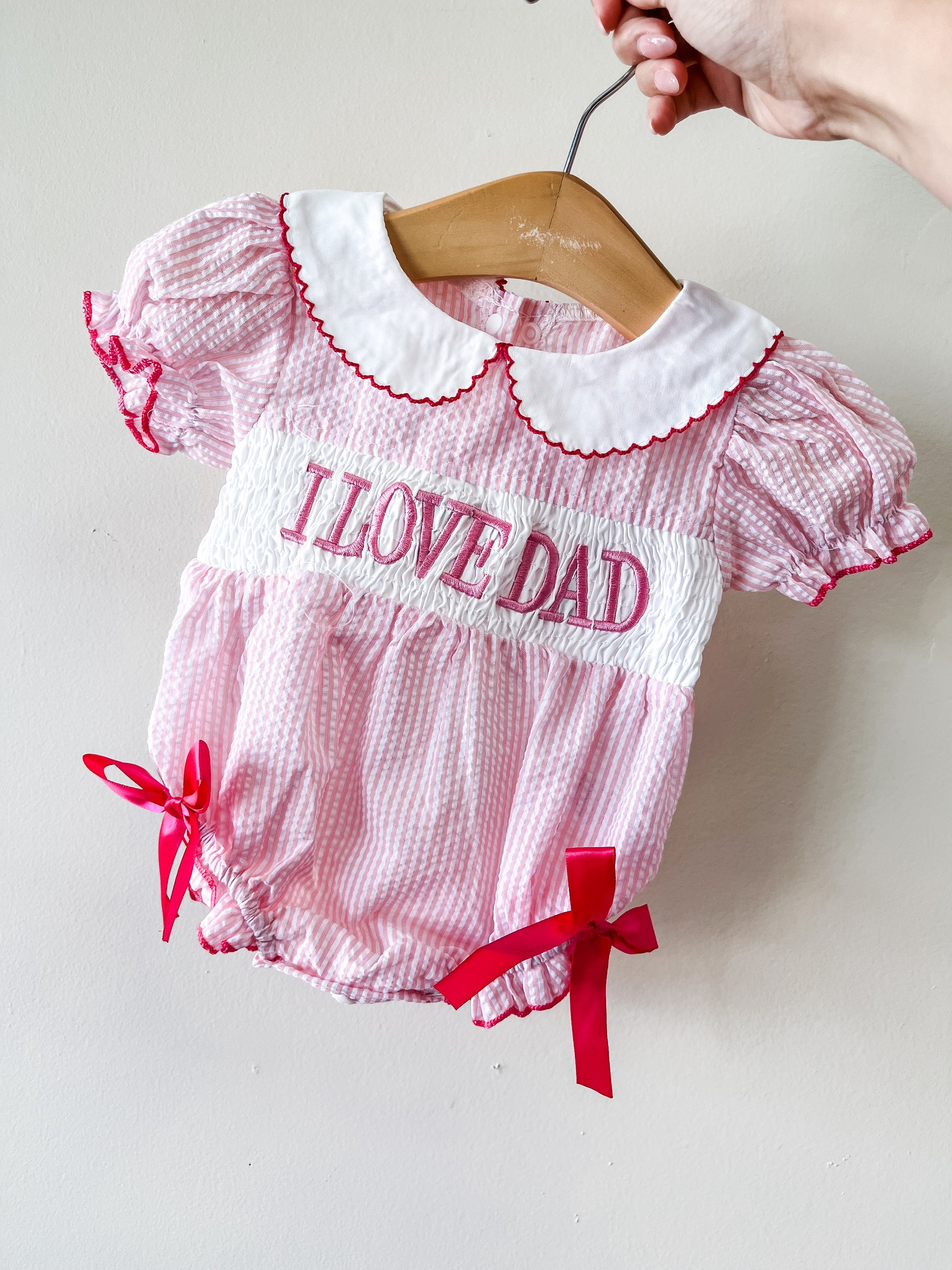 The Tiny Tot Bundles "I Love Dad Pink Bow Bubble" is a pink and white baby romper with puffed sleeves, a white Peter Pan collar, red trim, red leg bows, and "I LOVE DAD" in large red letters on the front. A hand holds it on a hanger.