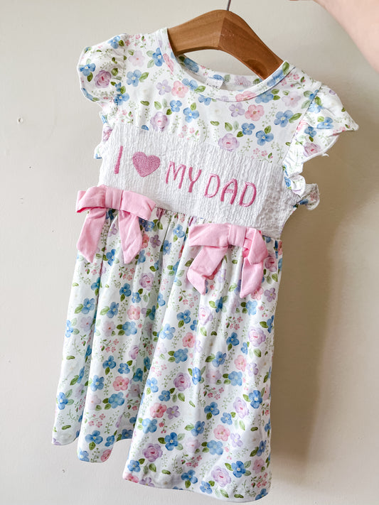 The Tiny Tot Bundles I Love Dad Floral Dress features pink bows, ruffled sleeves, and a white front panel embroidered with "I ♥ MY DAD." It hangs on a wooden hanger.