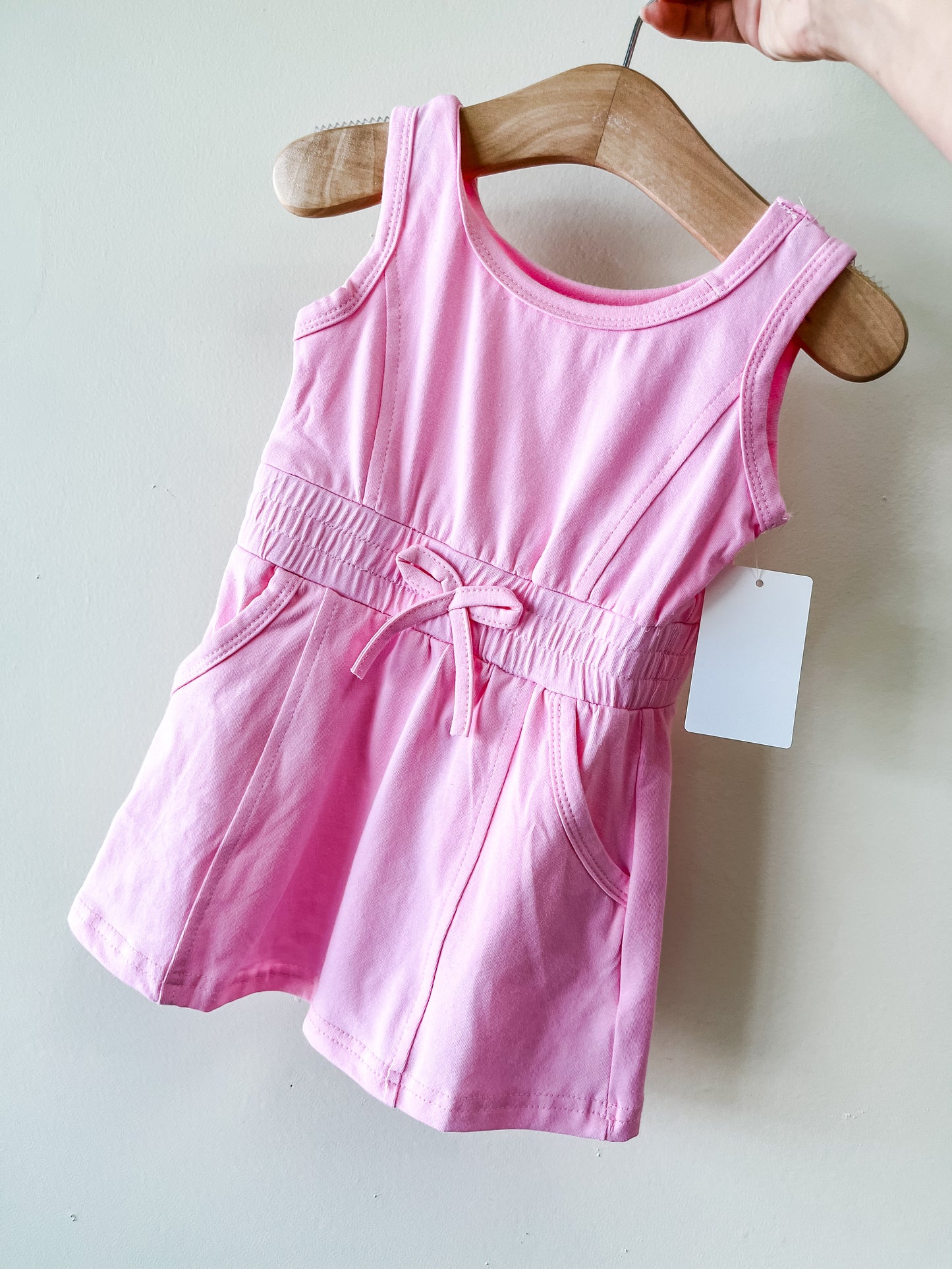 The Pink Sporty Dress by Tiny Tot Bundles is a sleeveless toddler dress with a gathered waist, front bow, and side pockets, shown on a wooden hanger with a blank tag.