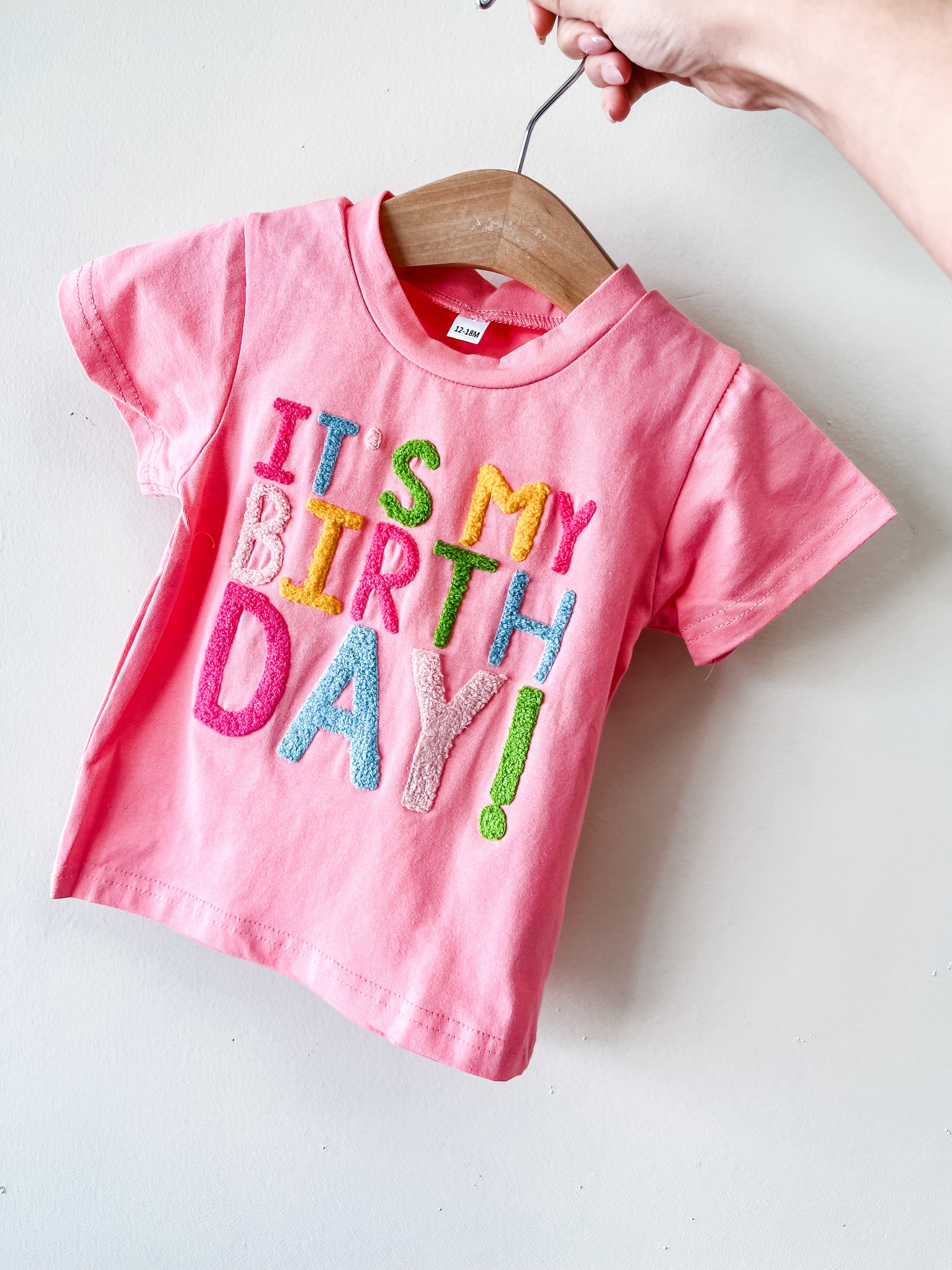 A hand holds a wooden hanger with Tiny Tot Bundles' It's My Birthday Tee—a pink toddler T-shirt featuring colorful, textured "ITS MY BIRTHDAY!" letters—against a white background.