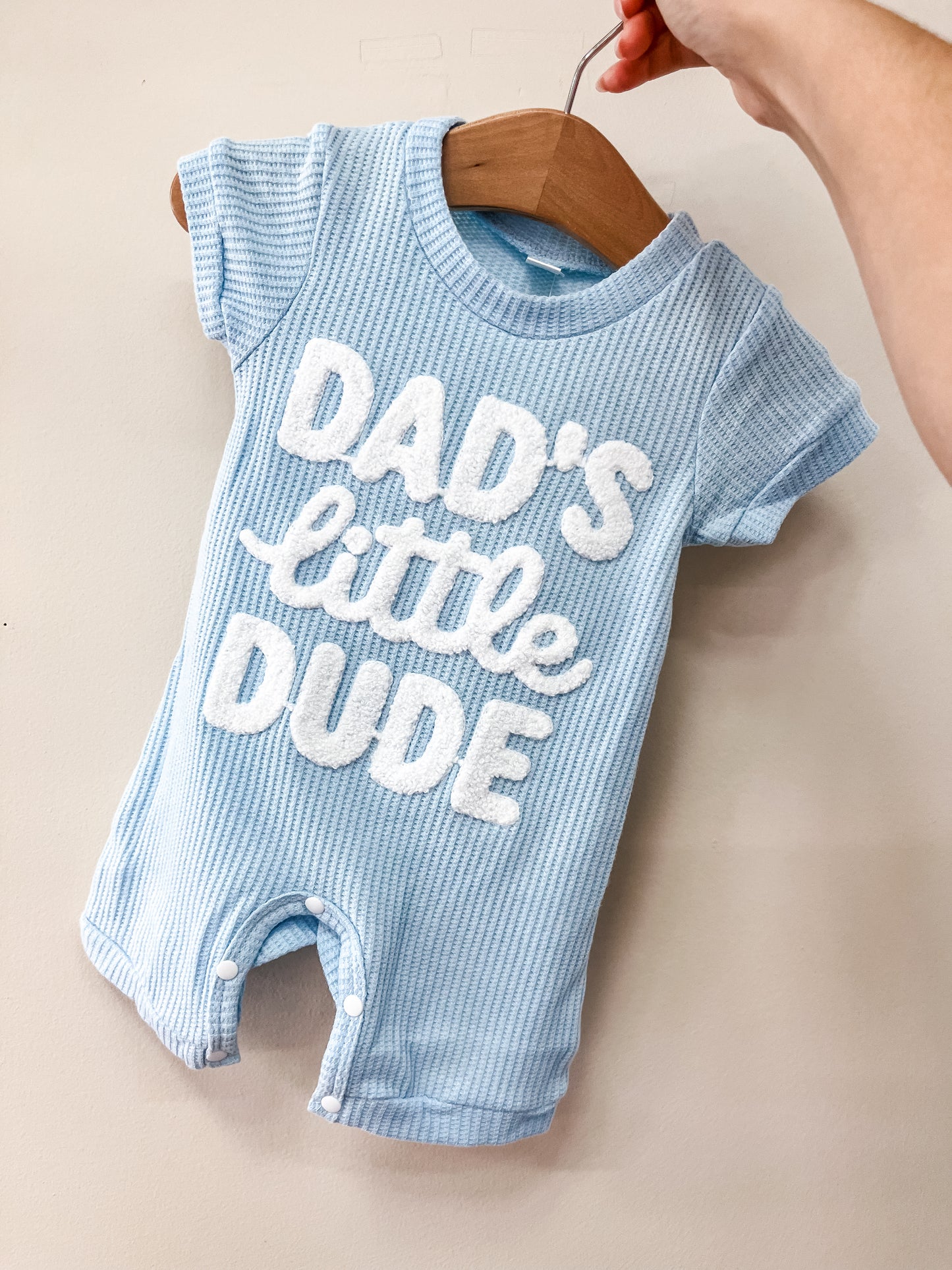 A hand holds a wooden hanger with the Tiny Tot Bundles Dad's Little Dude Romper, featuring short sleeves, snap buttons at the bottom, and "Dads little dude" in white textured letters on light blue fabric.