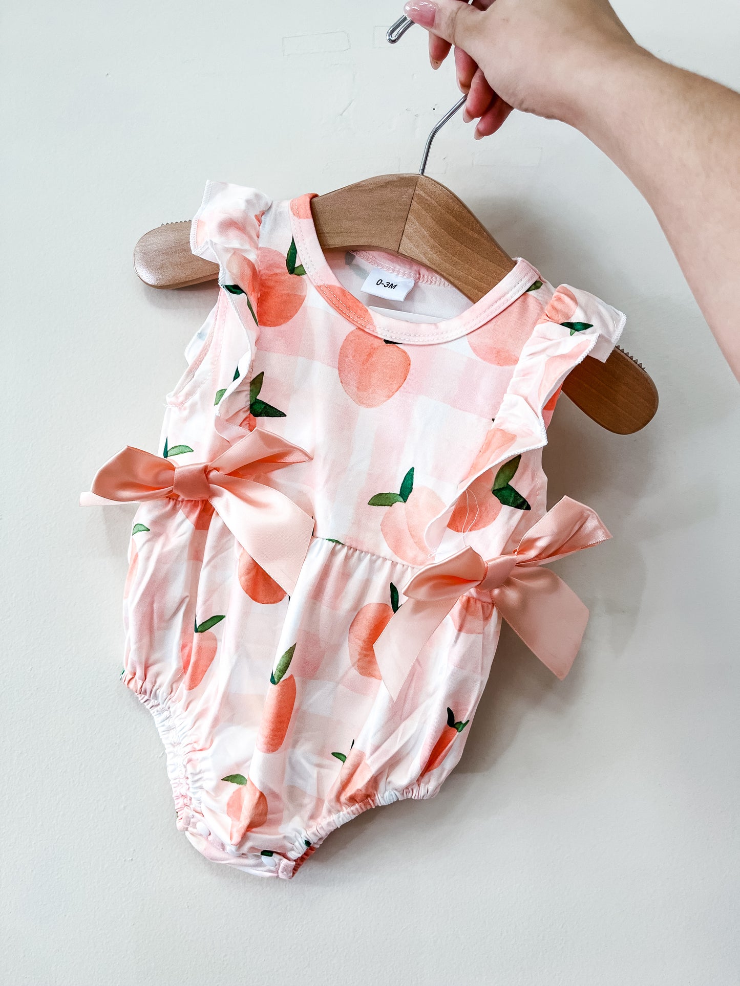 A hand holds the Tiny Tot Bundles Peach Bow Romper, featuring a peach print, ruffled sleeves, and two pink side bows, against a plain white background.