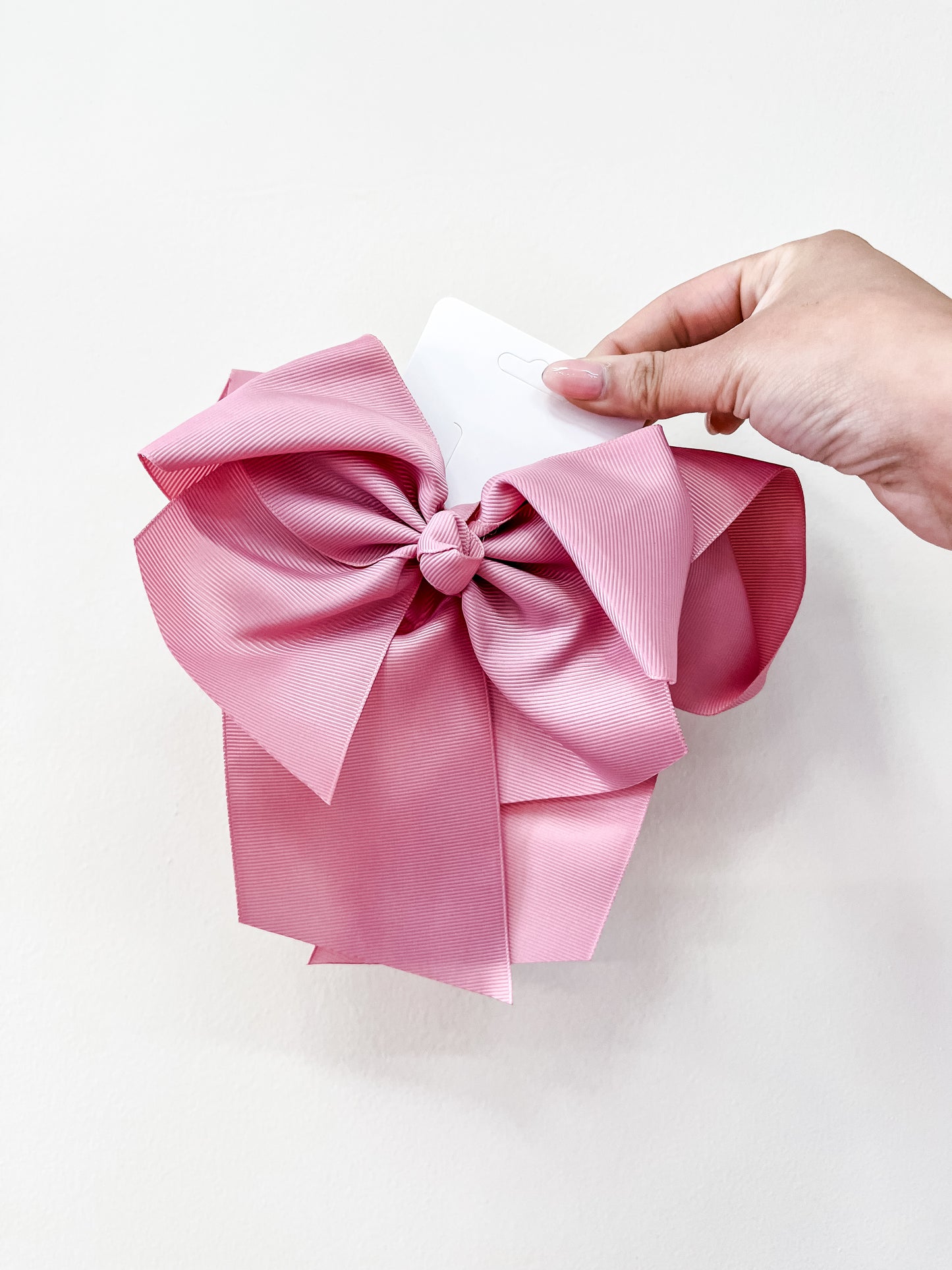 A hand holds the funteze Classic Statement Bow, a large pink ribbon with multiple loops and tails, against a plain white background.