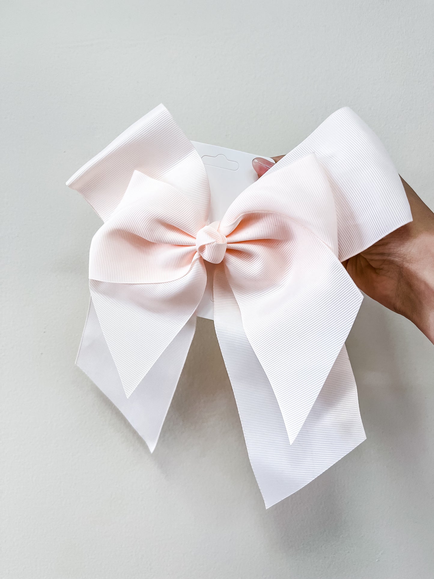 A hand holds the funteze Classic Statement Bow—a large, light pink accessory with wide, textured ribbons—against a plain white background.