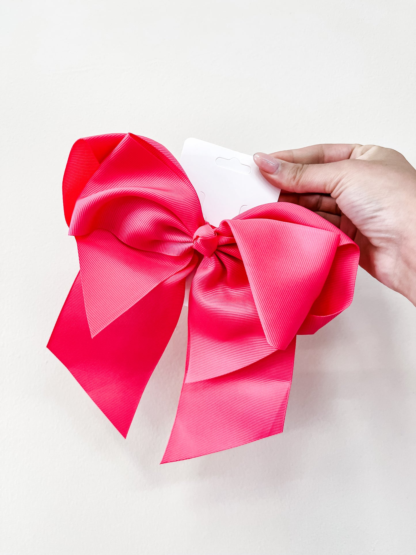 A hand holds the funteze Classic Statement Bow, a large, bright pink ribbon with two long tails, against a plain white background.