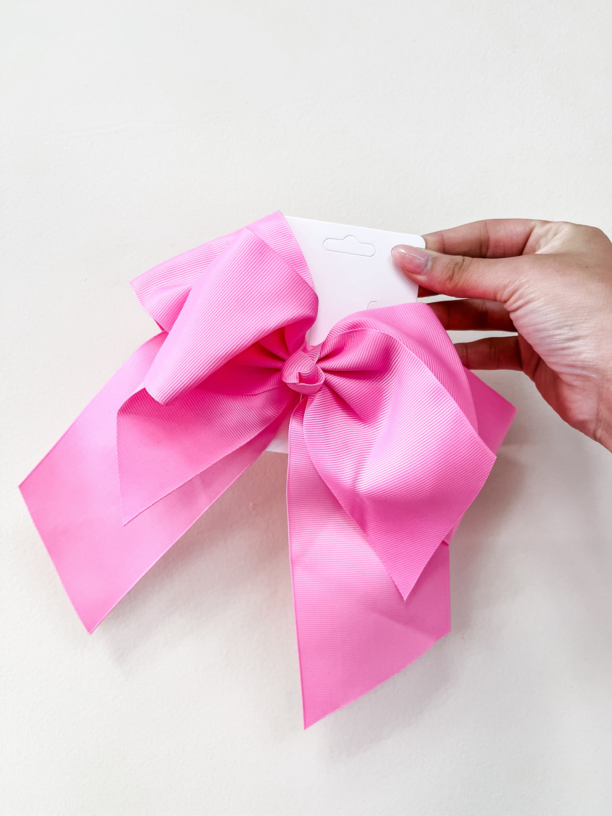 A hand holds the funteze Classic Statement Bow, a large, bright pink hair accessory attached to a white card, set against a plain, light-colored background.