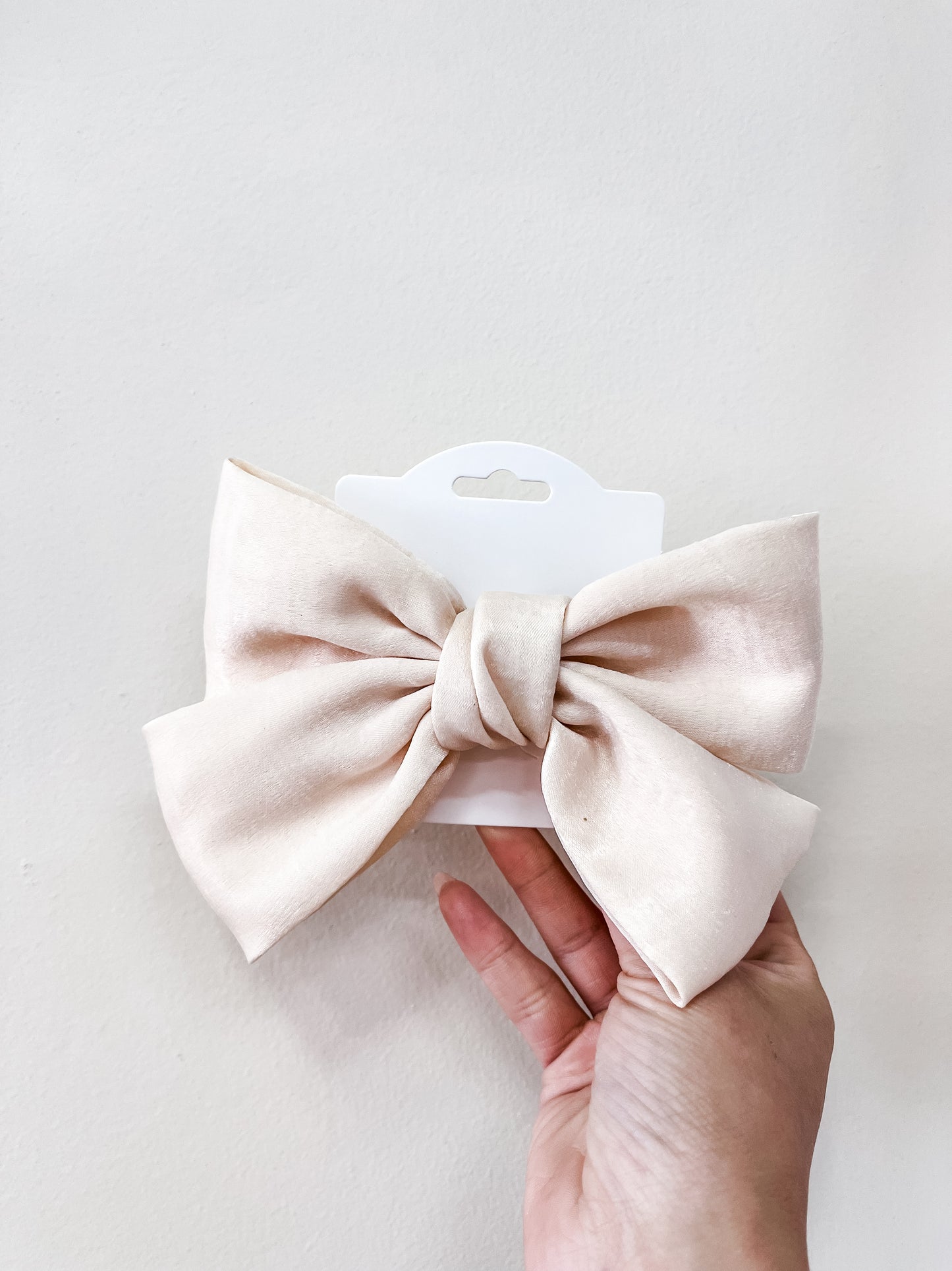 A hand holds the Luxe Velvet Statement Bow by Tiny Tot Bundles, in light beige, attached to a white display card against a plain off-white background.
