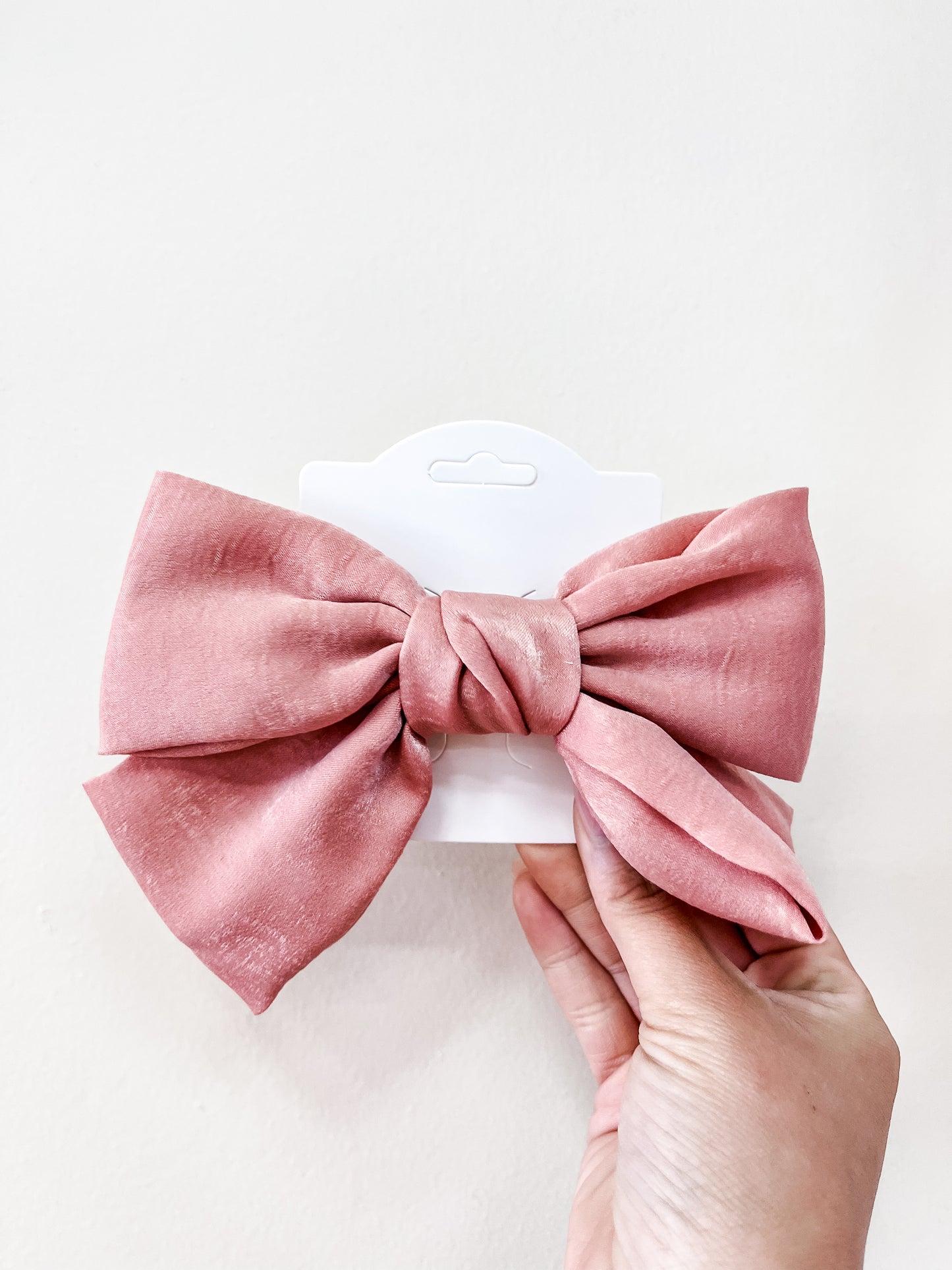 A hand holds the Luxe Velvet Statement Bow by Tiny Tot Bundles, a large pink fabric bow displayed on a white card against a plain light background.