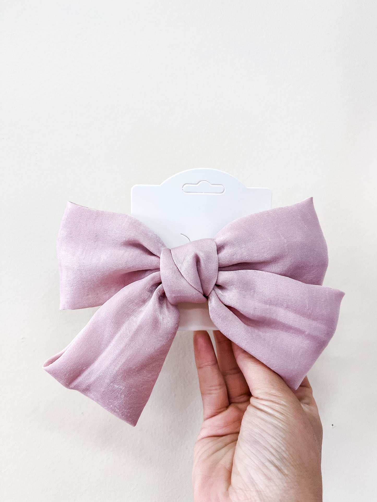 A hand holds the Luxe Velvet Statement Bow by Tiny Tot Bundles, a large light pink fabric bow attached to a white display card, set against a plain light background.