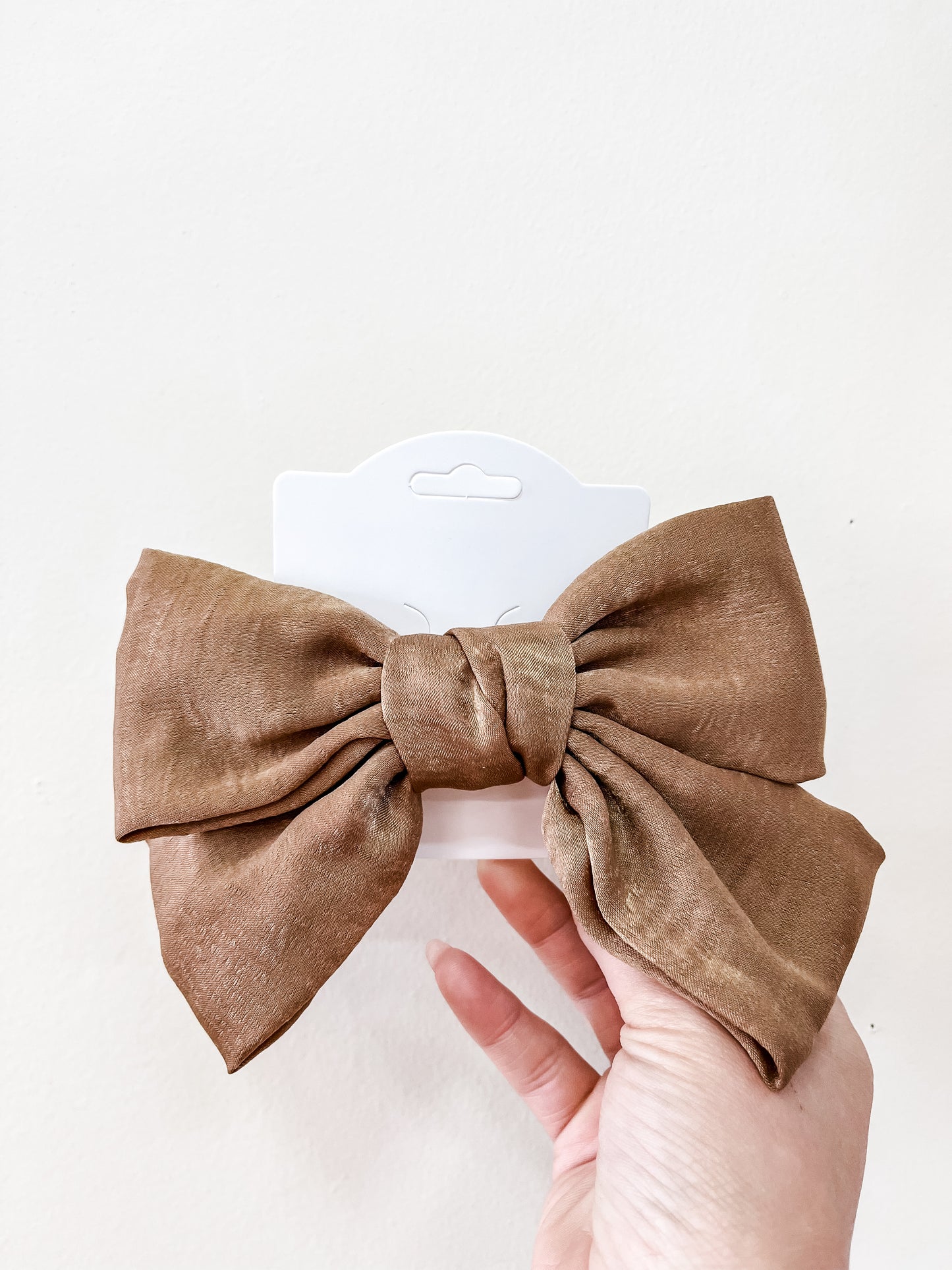 A hand holds the Luxe Velvet Statement Bow by Tiny Tot Bundles, a large brown velvet bow on a white display card, set against a plain light background.