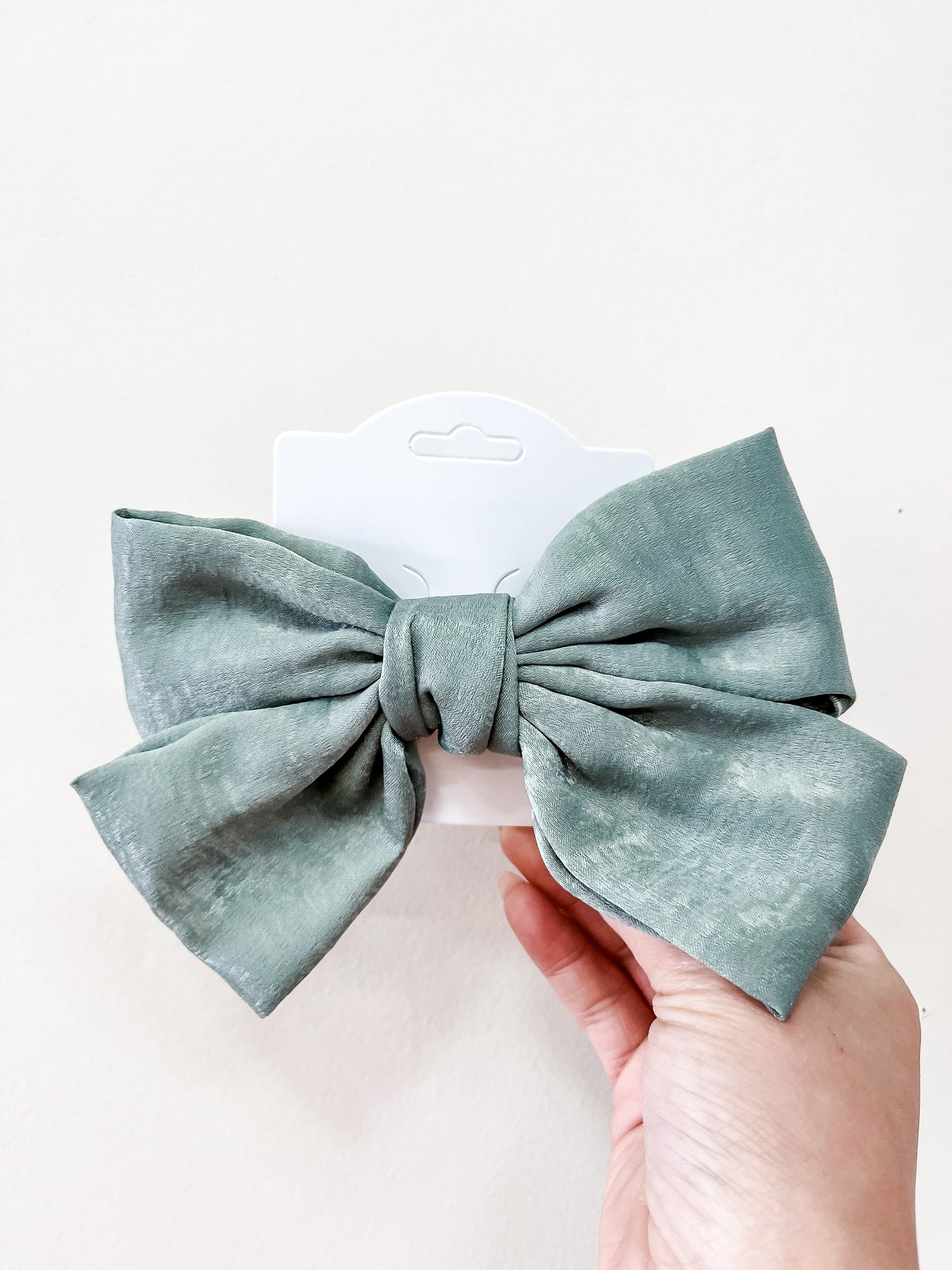 A hand holds the Luxe Velvet Statement Bow by Tiny Tot Bundles, a large pale green fabric bow displayed on a white card against a plain white background.