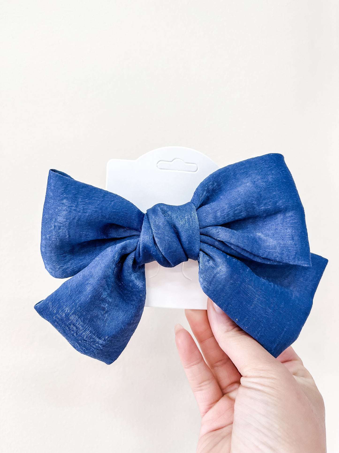 A hand holds the Luxe Velvet Statement Bow by Tiny Tot Bundles—a large blue velvet bow on a white display card—against a plain, light background.