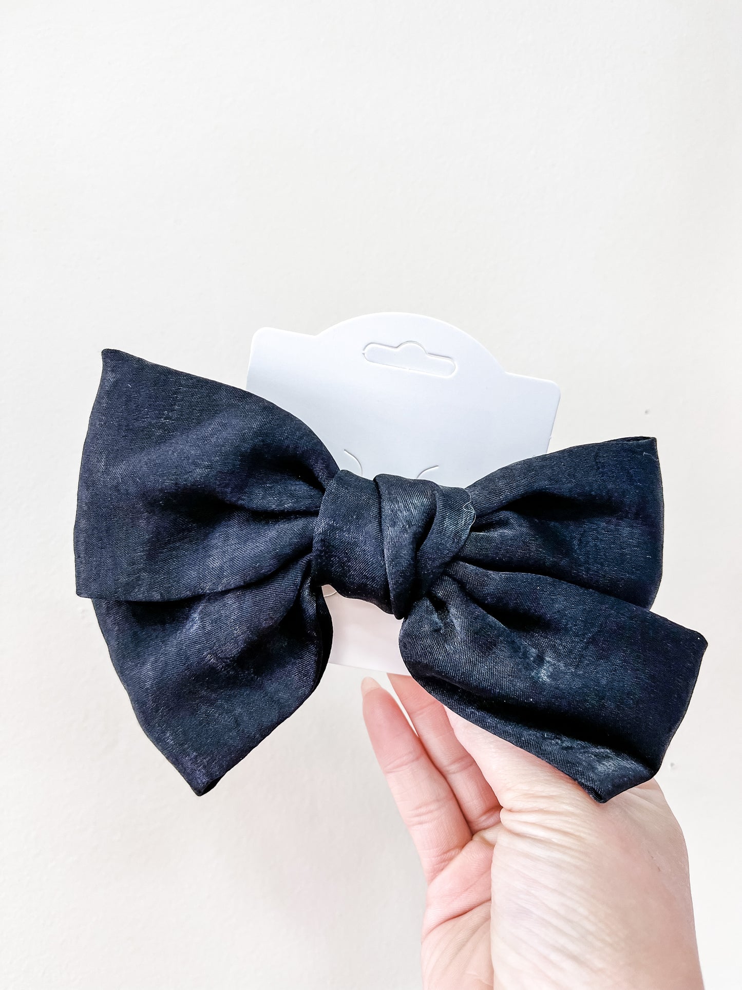 A hand holds the Luxe Velvet Statement Bow by Tiny Tot Bundles, a large black velvet bow attached to a white display card, set against a plain white background.