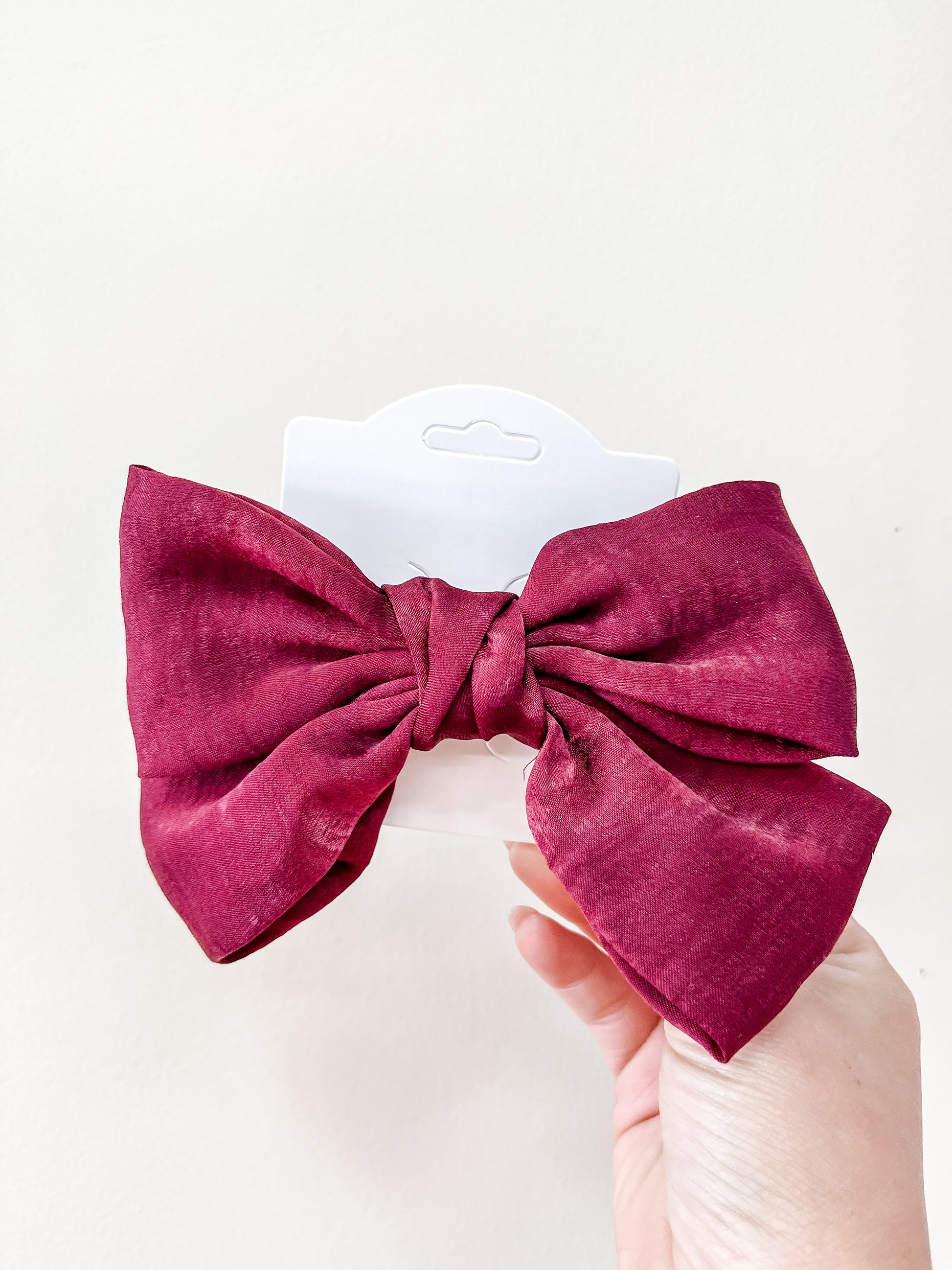 A hand holds the Luxe Velvet Statement Bow by Tiny Tot Bundles—a large, deep red bow on a white display card against a plain light background.