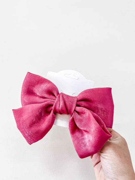 A hand holds the Luxe Velvet Statement Bow by Tiny Tot Bundles, a vibrant pink fabric hair bow displayed on white packaging against a plain light background.