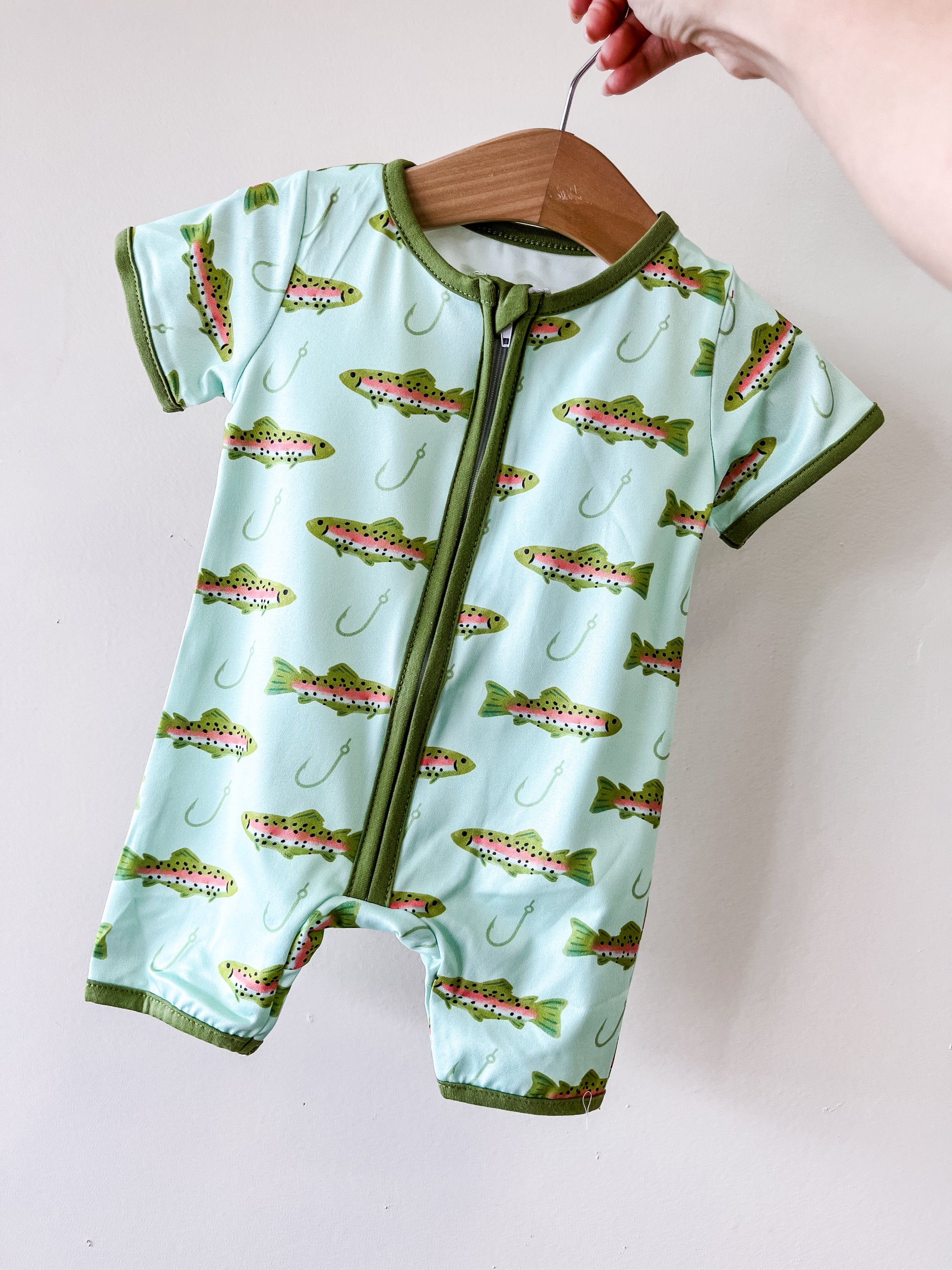 A hand holds up the Little Bass Buddy Romper by Tiny Tot Bundles—a light blue romper with green trim, fish and fishing hook pattern, and a front zipper—displayed on a wooden hanger.