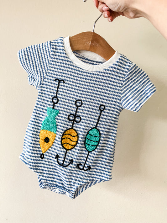 A hand holds up the Reelin’ in Summer Bubble by Tiny Tot Bundles, a blue-and-white striped baby romper with three colorful fishing lure embroideries on the front, displayed on a wooden hanger.