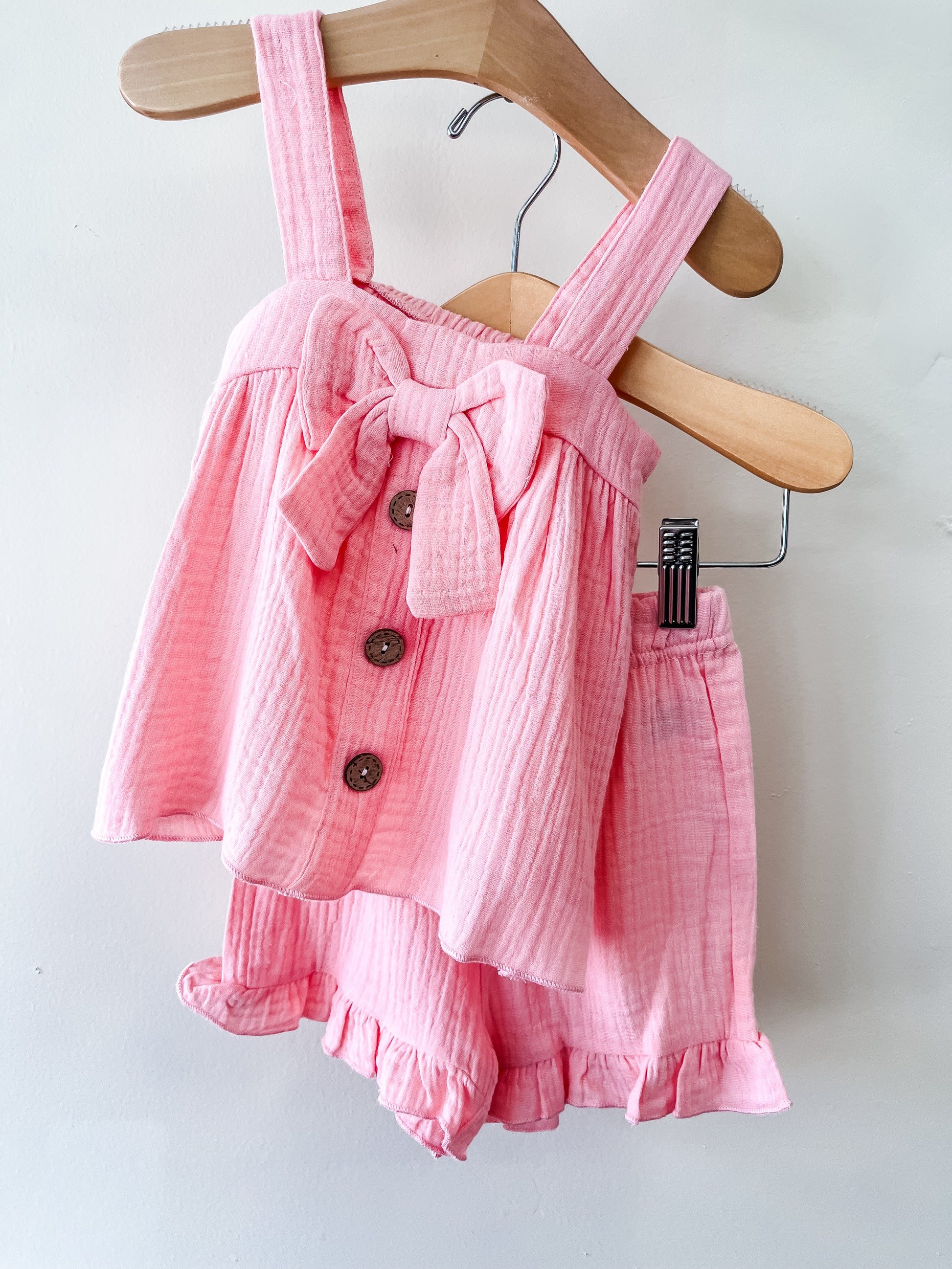 The Tiny Tot Bundles Sweet Peony Ruffle Set is a pink toddler girls’ outfit with a sleeveless top featuring thick straps, a large bow, three buttons, and matching ruffled hem shorts, displayed on a hanger.