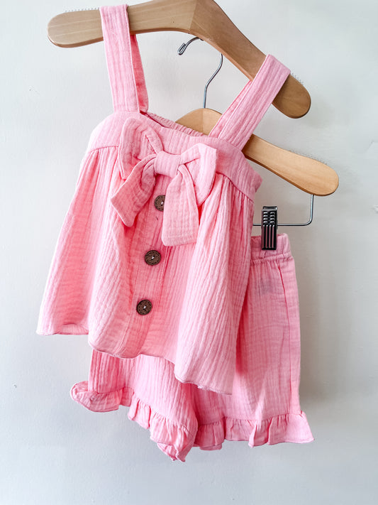 The Tiny Tot Bundles Sweet Peony Ruffle Set is a pink toddler girls’ outfit with a sleeveless top featuring thick straps, a large bow, three buttons, and matching ruffled hem shorts, displayed on a hanger.