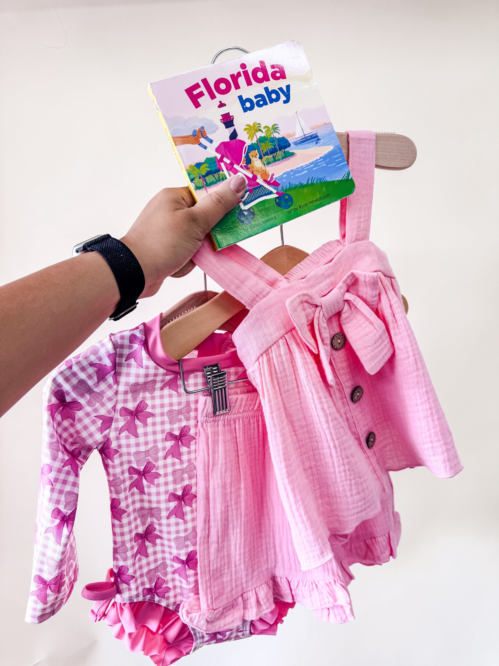 A hand holds the Coastal Coquette Bundle by Tiny Tot Bundles above two pink toddler outfits: a gingham swimsuit with bows and a sleeveless dress with buttons and a front bow.