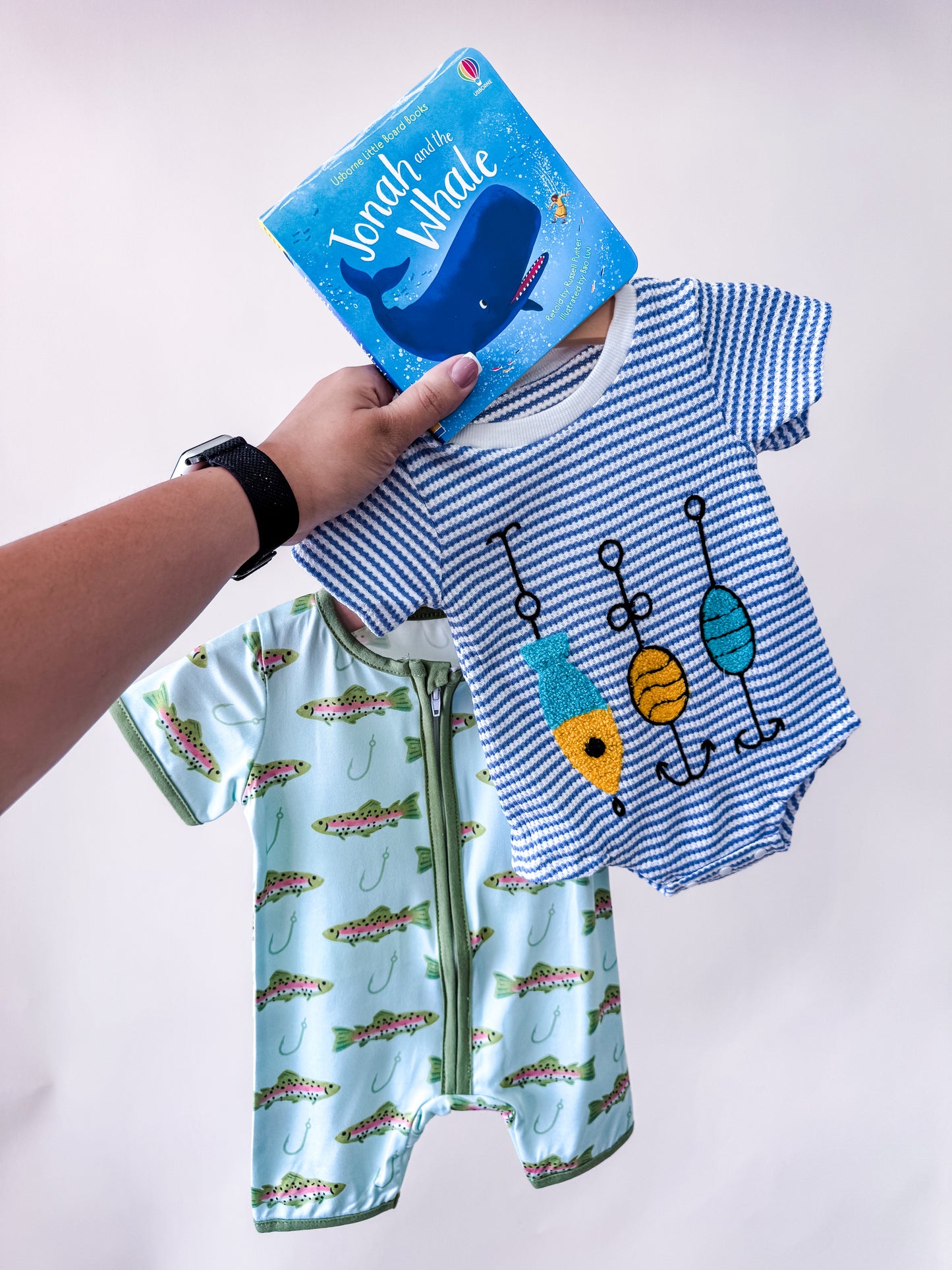 A hand holds the Tiny Tot Bundles Reel Cute Boy Bundle, featuring a blue "Jonah and the Whale" children’s book and two baby outfits: one with blue-and-white stripes and fish, and one green with alligators, all set against a plain background.