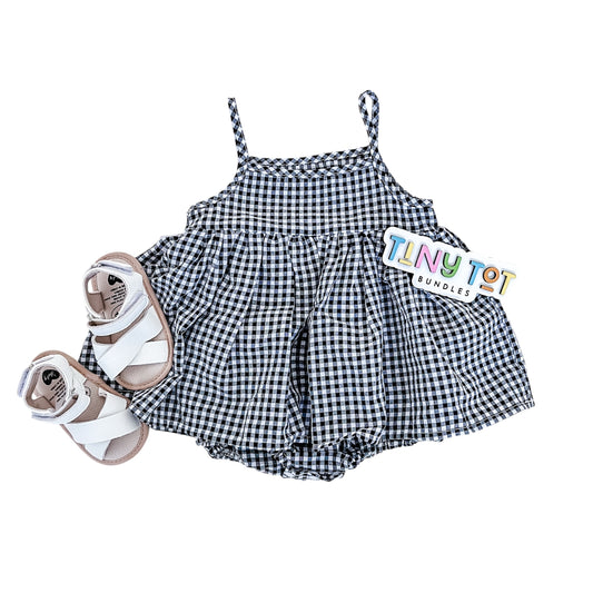 The Natalia Romper by Tiny Tot Bundles, a black and white gingham baby dress with matching bloomers and white sandals, is displayed with a colorful Tiny Tot Bundles tag on a white background.