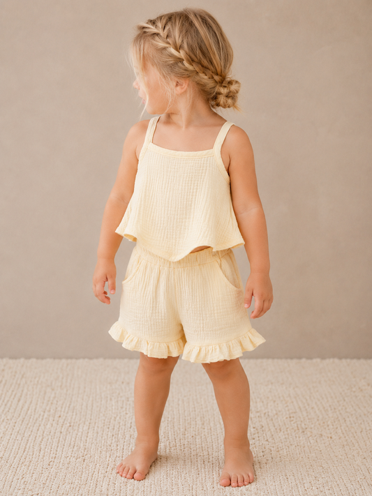 A young child stands barefoot on a beige rug, wearing the Dainty Sunshine Set by Tiny Tot Bundles: a light yellow, sleeveless top with matching ruffled shorts; her blonde hair is styled in braids as she looks to the side against a neutral background.
