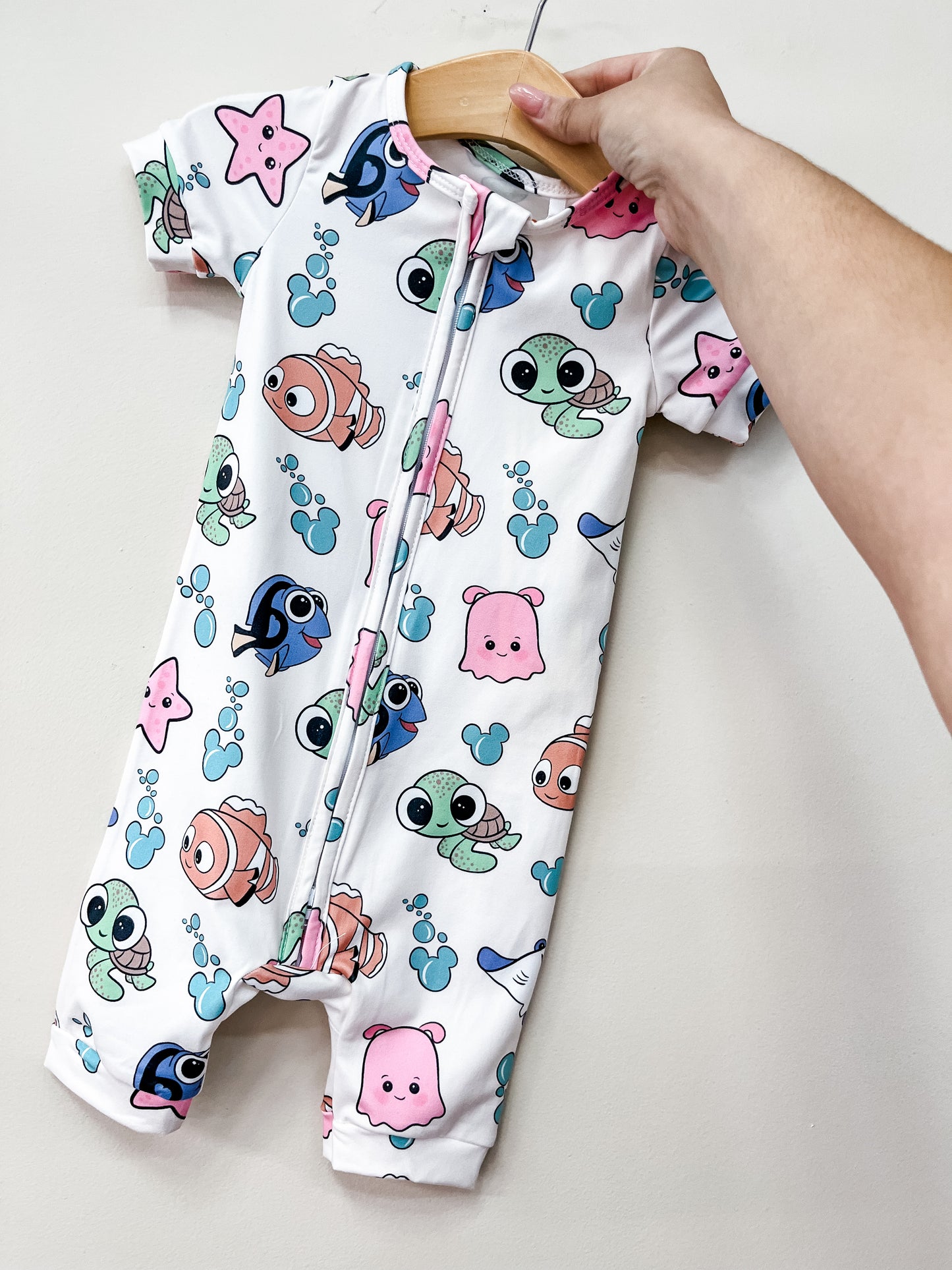 A hand displays the Tiny Tot Bundles Baby Nemo Shorty, a white romper with a front zipper, featuring colorful cartoon turtles, fish, octopuses, and starfish.