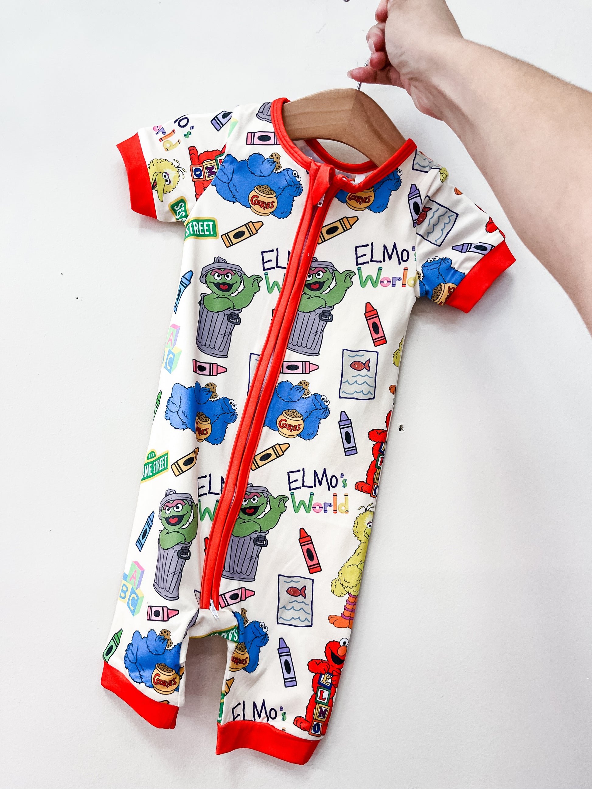 A hand holds up the Sesame Street Shorty by Tiny Tot Bundles, a baby romper featuring Elmo, Oscar the Grouch, Cookie Monster, Big Bird, colorful crayons, “Elmos World” text, red trim, and a convenient zipper.