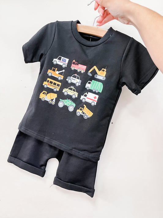 The Tiny Tot Bundles Transportation Crew Shorts Set is a black short-sleeve toddler outfit with matching shorts, featuring colorful vehicle illustrations like cars, trucks, buses, and construction equipment on the shirt. Displayed on a hanger.