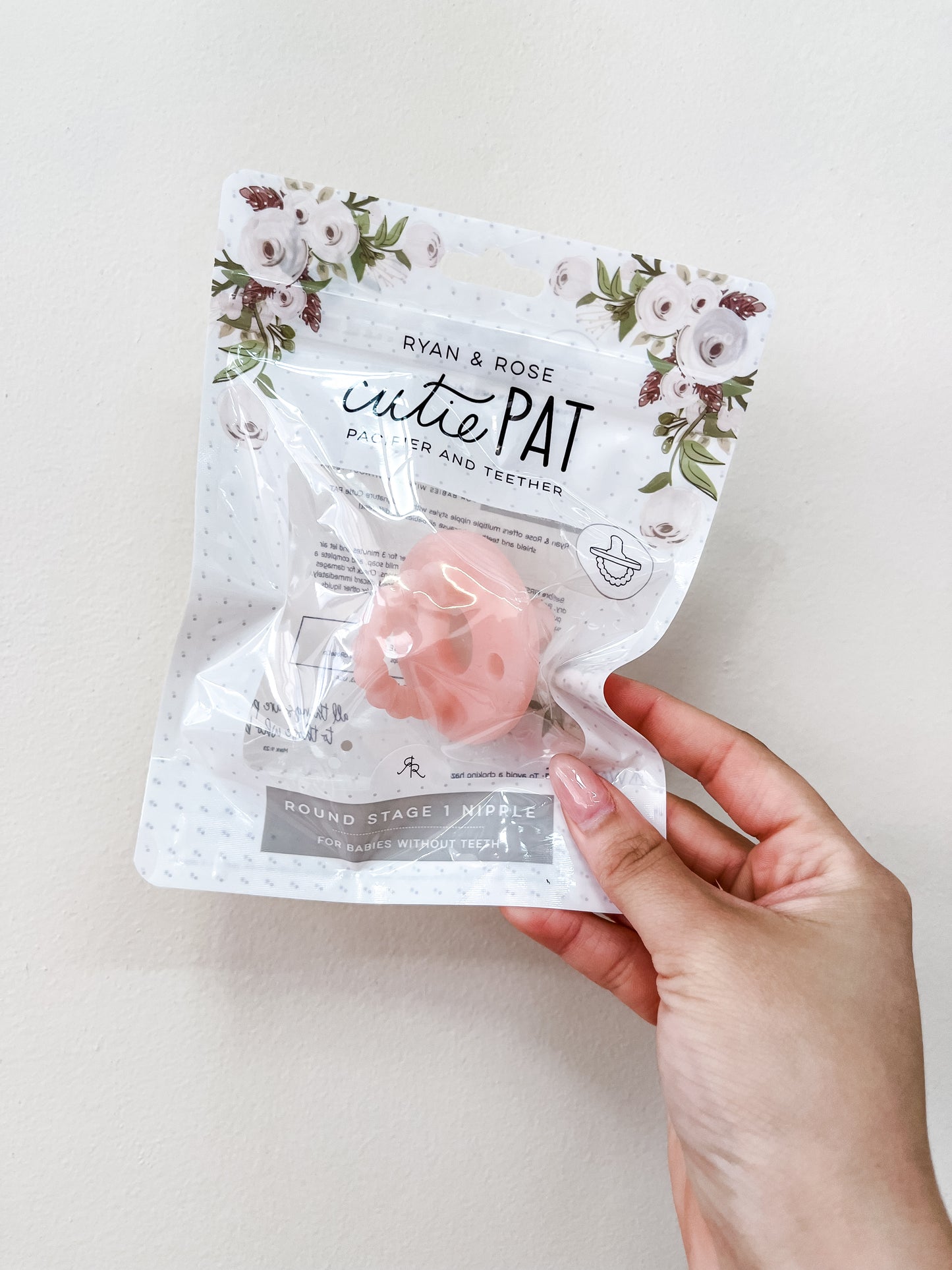 A hand holds a sealed package of the ryan & Rose Cutie Pat Pacifier - Clear Pink against a light background; the packaging features white flowers at the top.