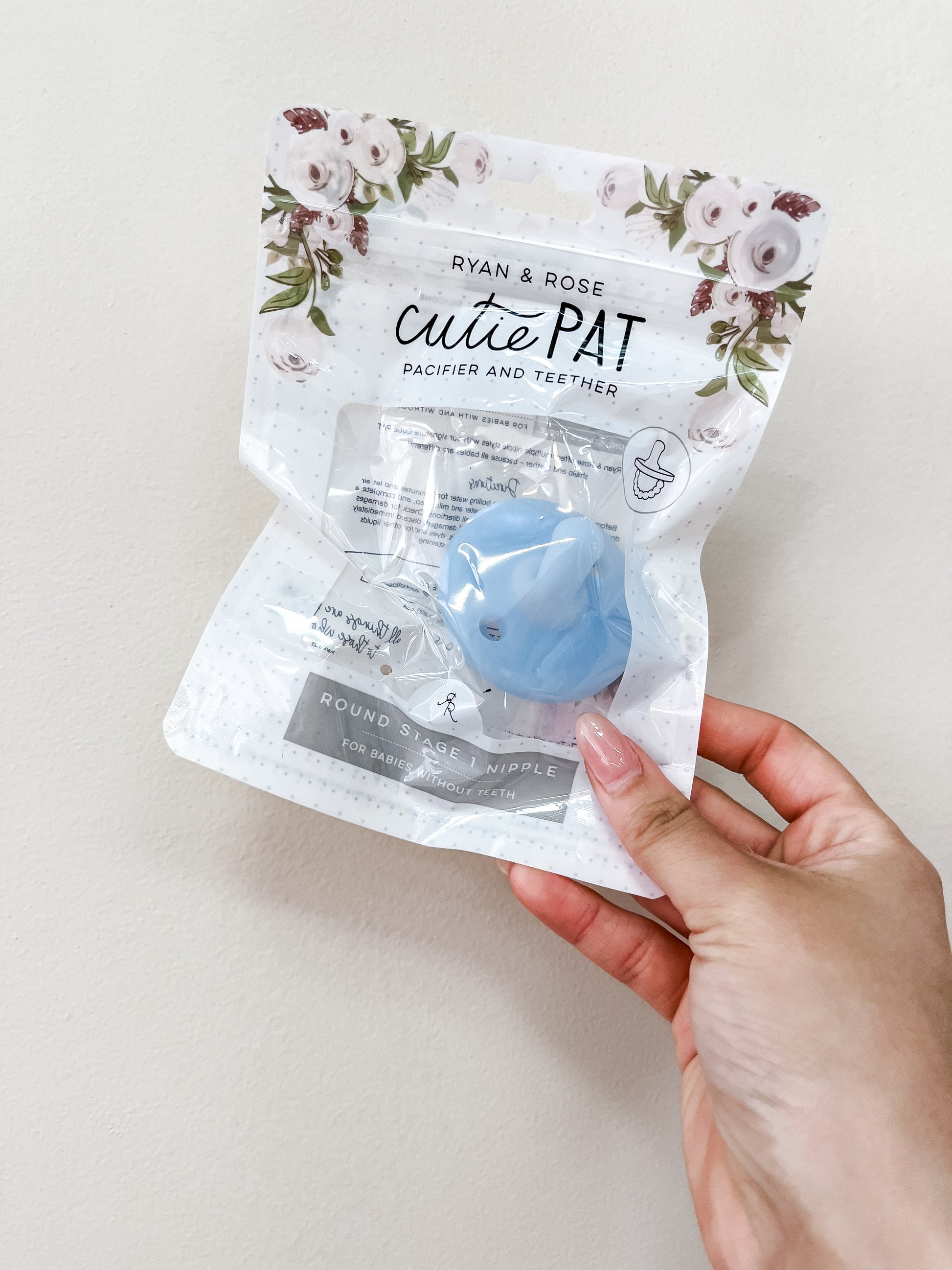 A hand holds a sealed package of the ryan & Rose Cutie Pat Pacifier - Clear Blue, with floral designs on the top.