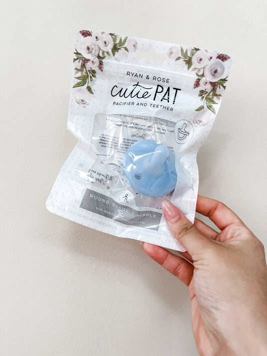 A hand holds a sealed package of the ryan & Rose Cutie Pat Pacifier - Clear Blue, with floral designs on the top.