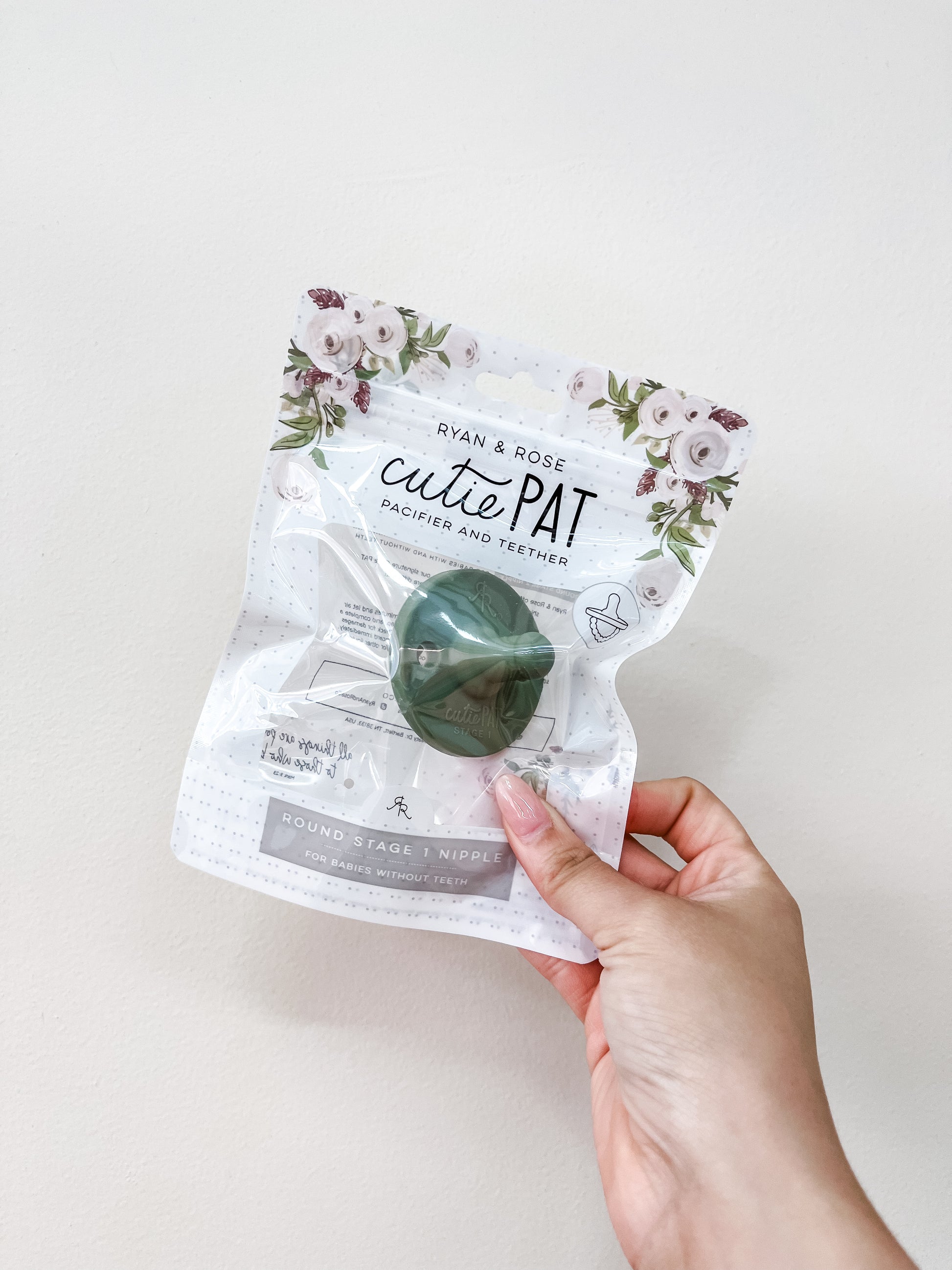 A hand holds a clear package of the ryan & Rose Cutie Pat Pacifier in Sage green, showcasing the floral design and green pacifier inside.