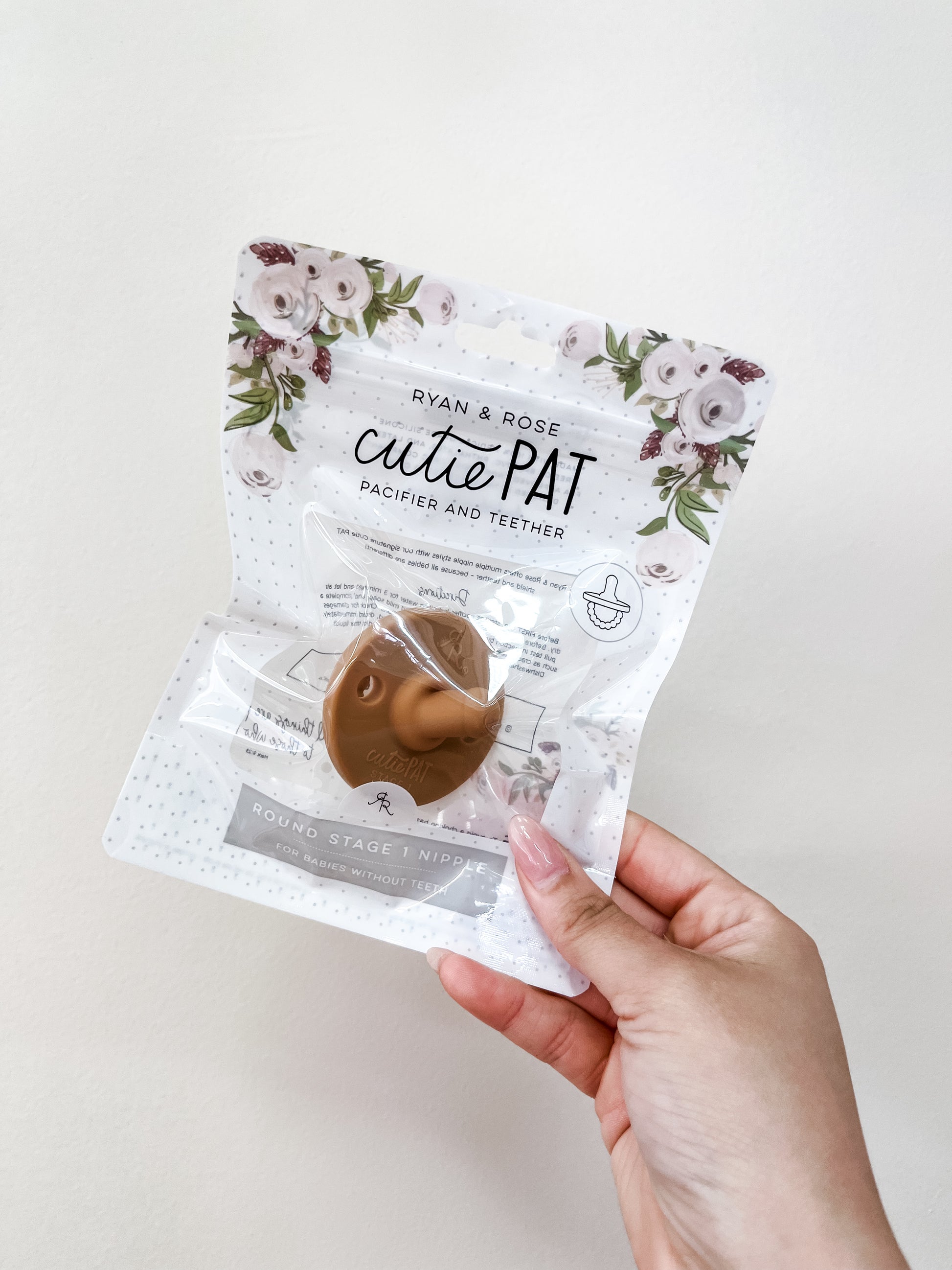 A hand holds a tan Cutie Pat Pacifier by ryan & Rose in clear packaging with the brand name and product name visible.