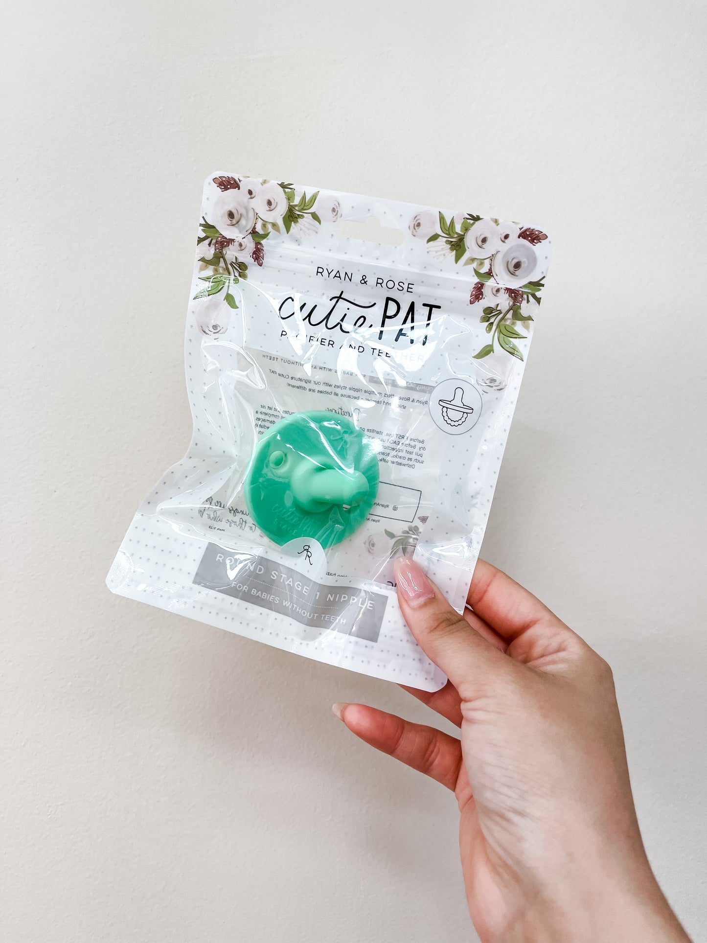 A hand holds a clear package labeled ryan & Rose Cutie Pat Pacifier - Green, displaying a mint green pacifier and floral designs on the top of the packaging.