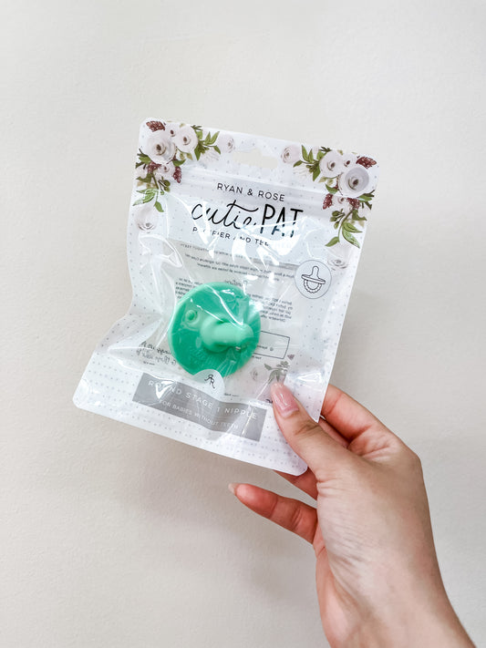 A hand holds a clear package labeled ryan & Rose Cutie Pat Pacifier - Green, displaying a mint green pacifier and floral designs on the top of the packaging.