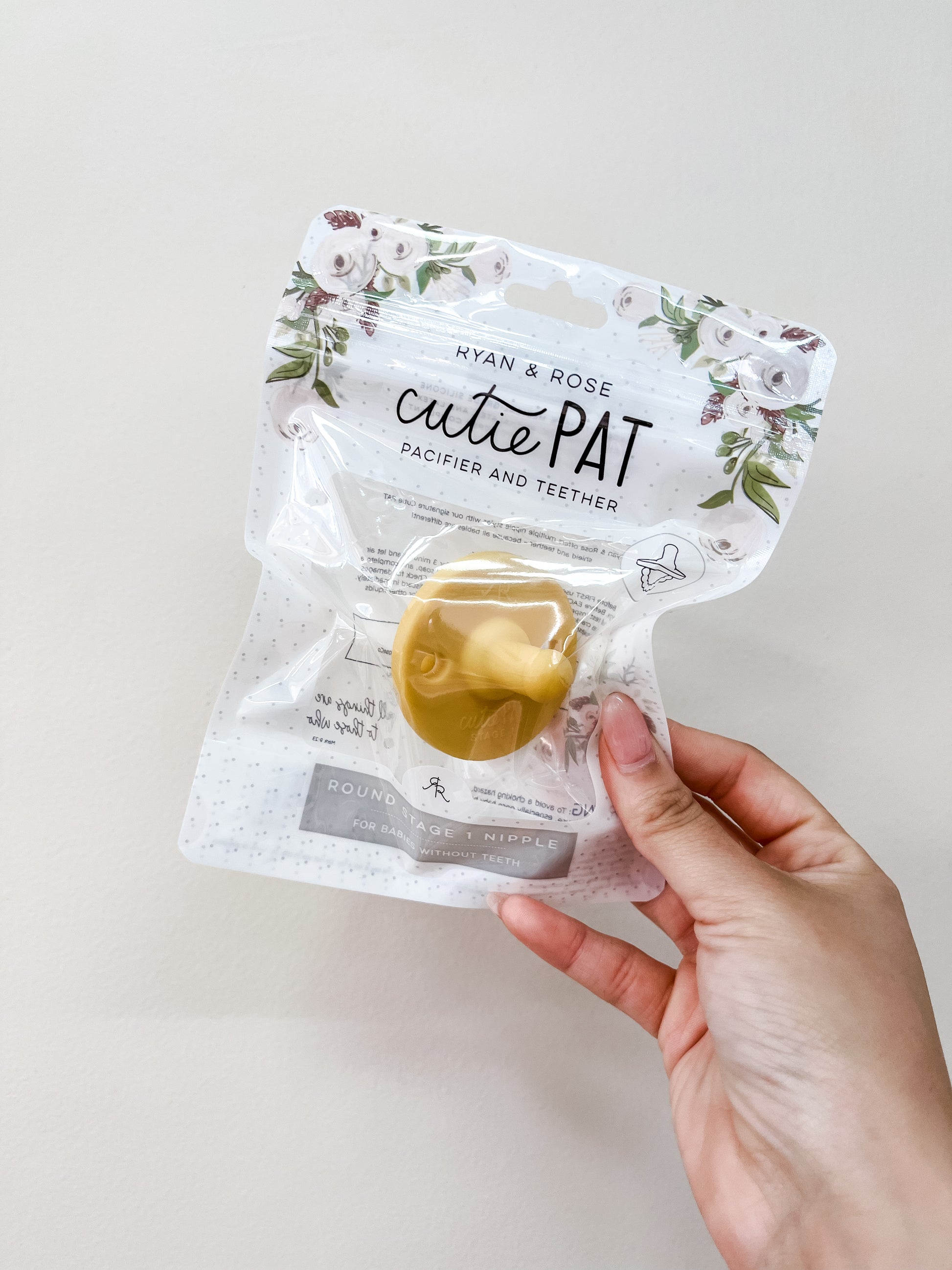 A hand holds a Cutie Pat Pacifier - Yellow by ryan & Rose in clear packaging with floral designs and product information.