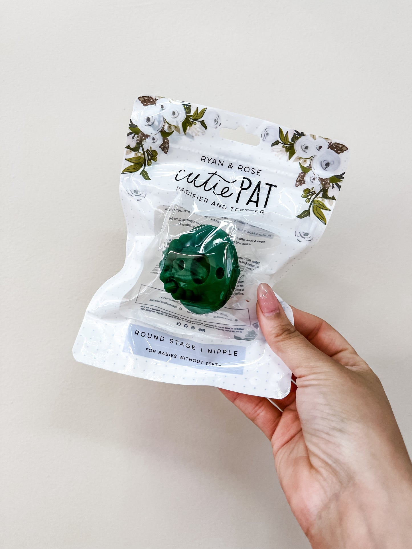 A hand holds a packaged Cutie Pat Pacifier - Forest Green by ryan & Rose, featuring floral designs on the packaging, against a plain light background.