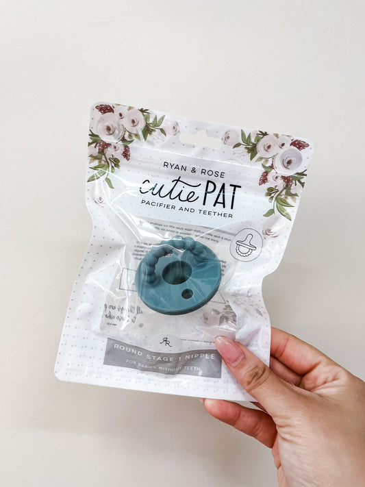 A hand holds a packaged sky blue Cutie Pat Pacifier by ryan & Rose, with floral designs on the packaging.