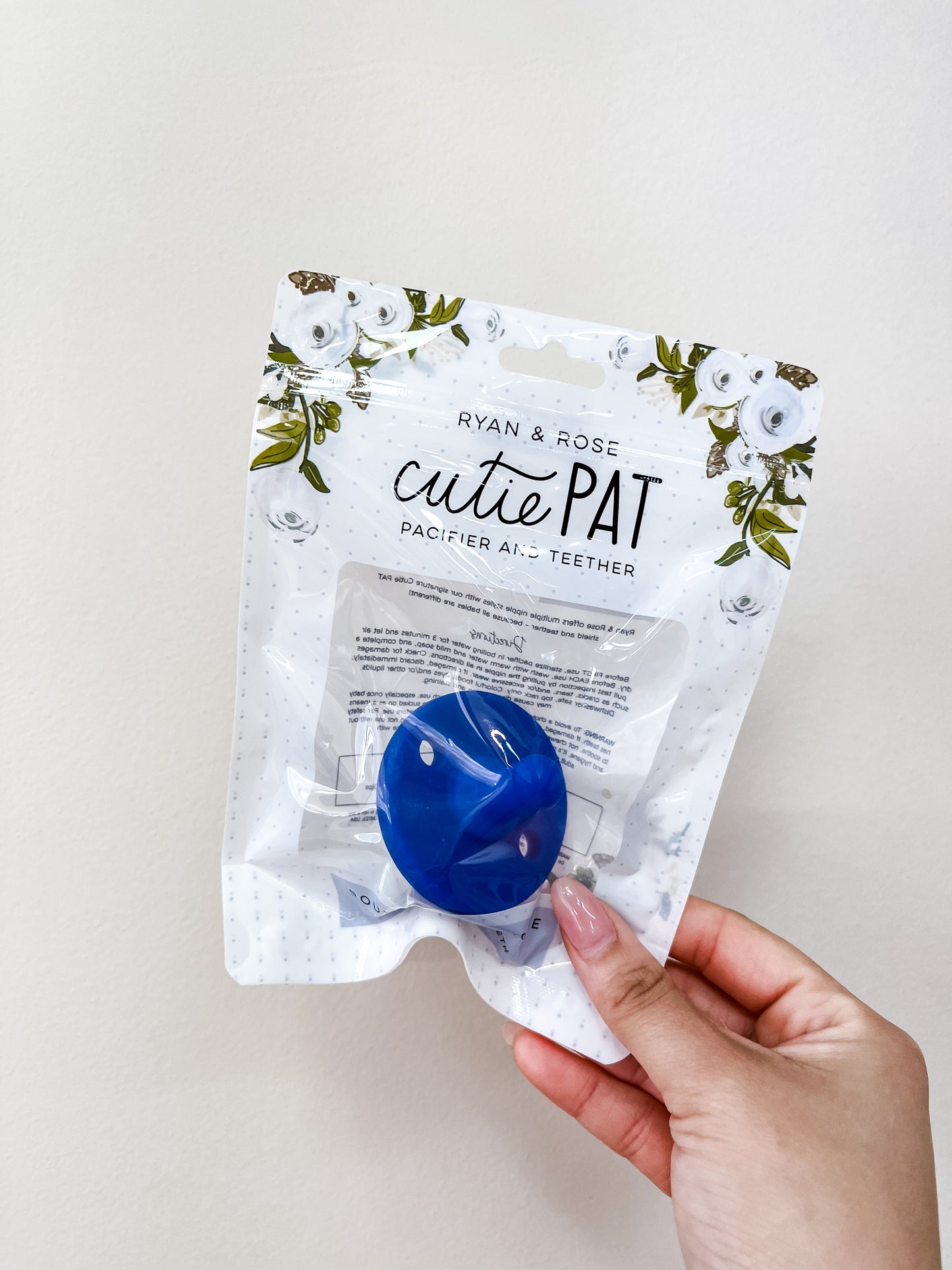 A hand holds a packaged Cutie Pat Pacifier - Royal Blue by ryan & Rose. The white packaging features floral designs and a clear plastic window displaying the blue pacifier inside.