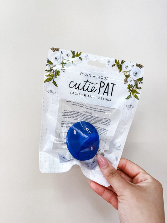 A hand holds a packaged Cutie Pat Pacifier - Royal Blue by ryan & Rose. The white packaging features floral designs and a clear plastic window displaying the blue pacifier inside.