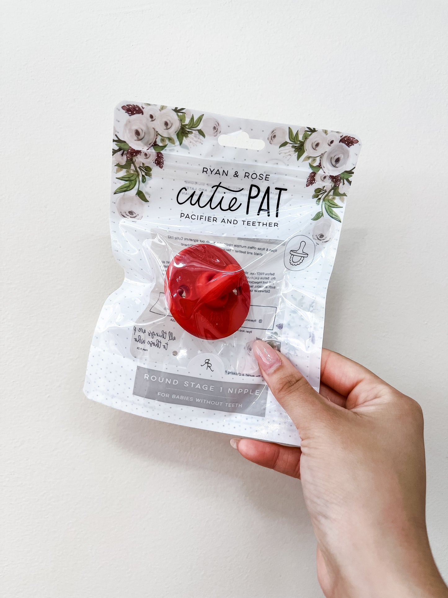 A hand holds a packaged Cutie Pat Pacifier - Red by ryan & Rose with floral decorations on the packaging, set against a plain white background.