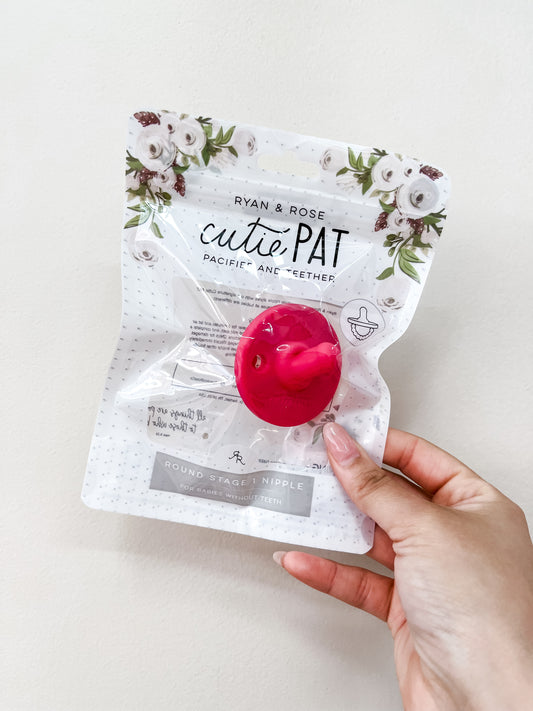 A hand holds a ryan & Rose Cutie Pat Pacifier - Hot Pink, visible in its floral-topped package against a plain white background.