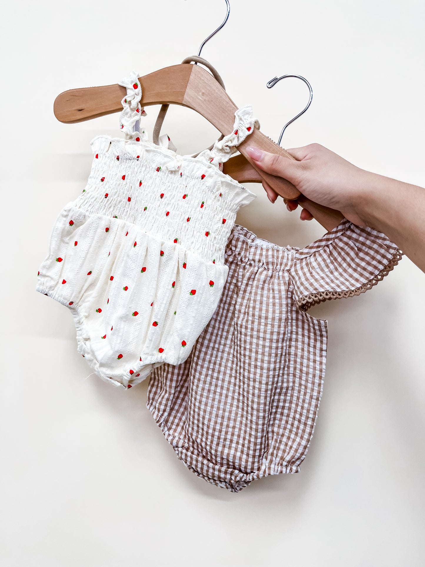 A hand displays the Tiny Tot Bundles "Bundle Of The Day - Girls": two baby rompers—one white with red polka dots and smocking, the other brown and white gingham with puffed sleeves—on wooden hangers against a plain white background.